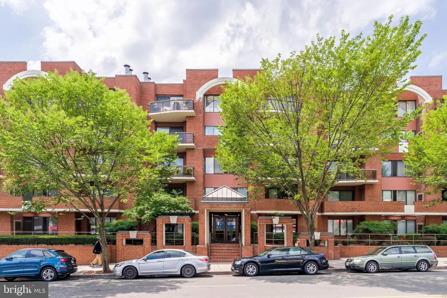 A brick apartment building with green trees in front and four parked cars along the street.