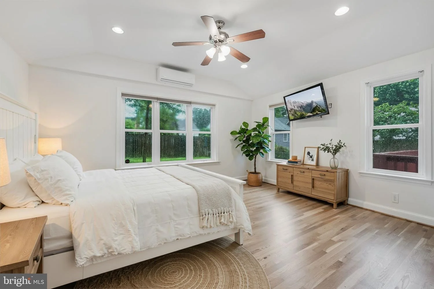 Bright bedroom with white bedding, wooden furniture, hardwood floors, windows showing green outdoor scenery, ceiling fan, wall-mounted TV, potted plant, and decorative items on a wooden dresser.