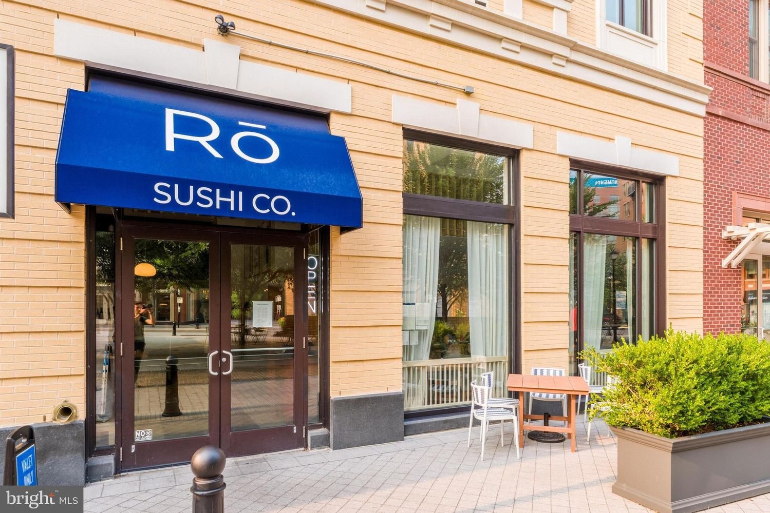 Restaurant with a blue awning reading 'RO SUSHI CO.' in front of sidewalk seating with small table and chairs, large windows, and a brick building exterior.