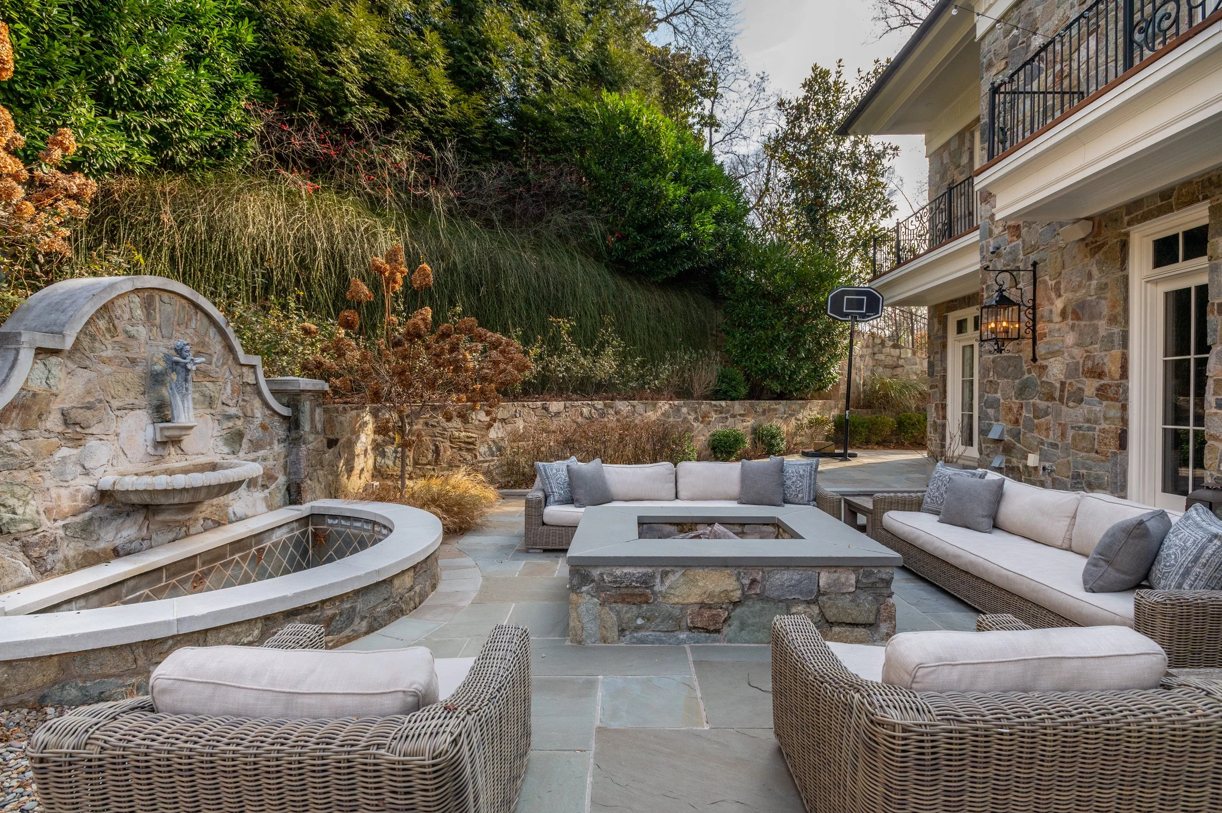 An outdoor patio area with a stone wall fountain and fountain basin, outdoor seating with cushions, and a basketball hoop, surrounded by greenery and a stone building with balconies.