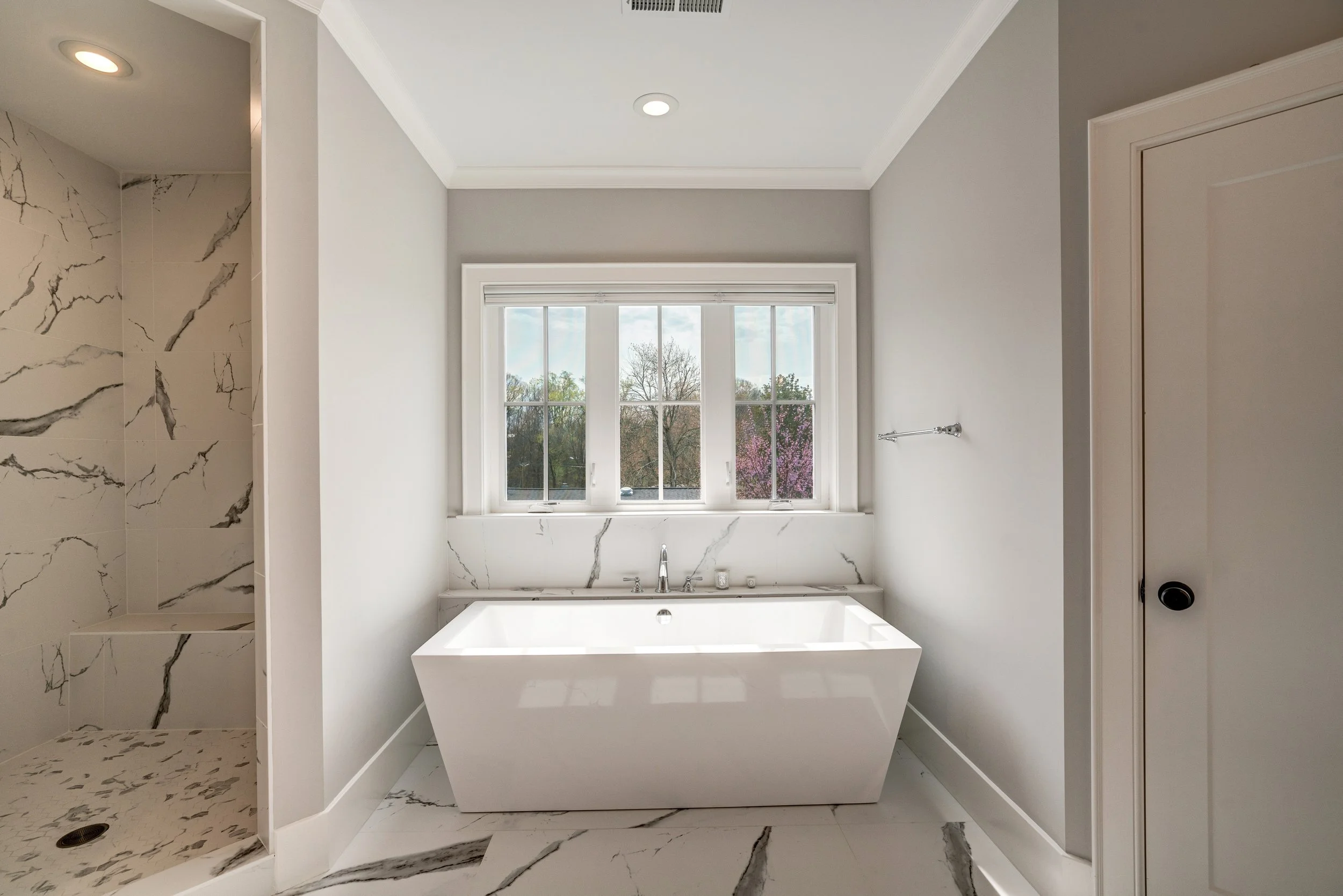 Modern bathroom with large bathtub beneath a window, marble-tiled shower area to the left, and neutral color scheme.