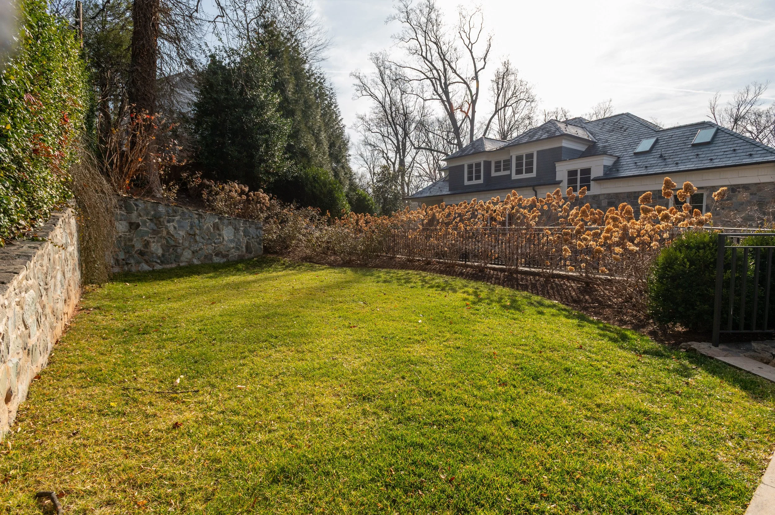 A backyard lawn with a stone retaining wall on the left, a wrought iron fence with dried ornamental grasses, a two-story house with a slate roof, and tall leafless trees in the background under a partly cloudy sky.