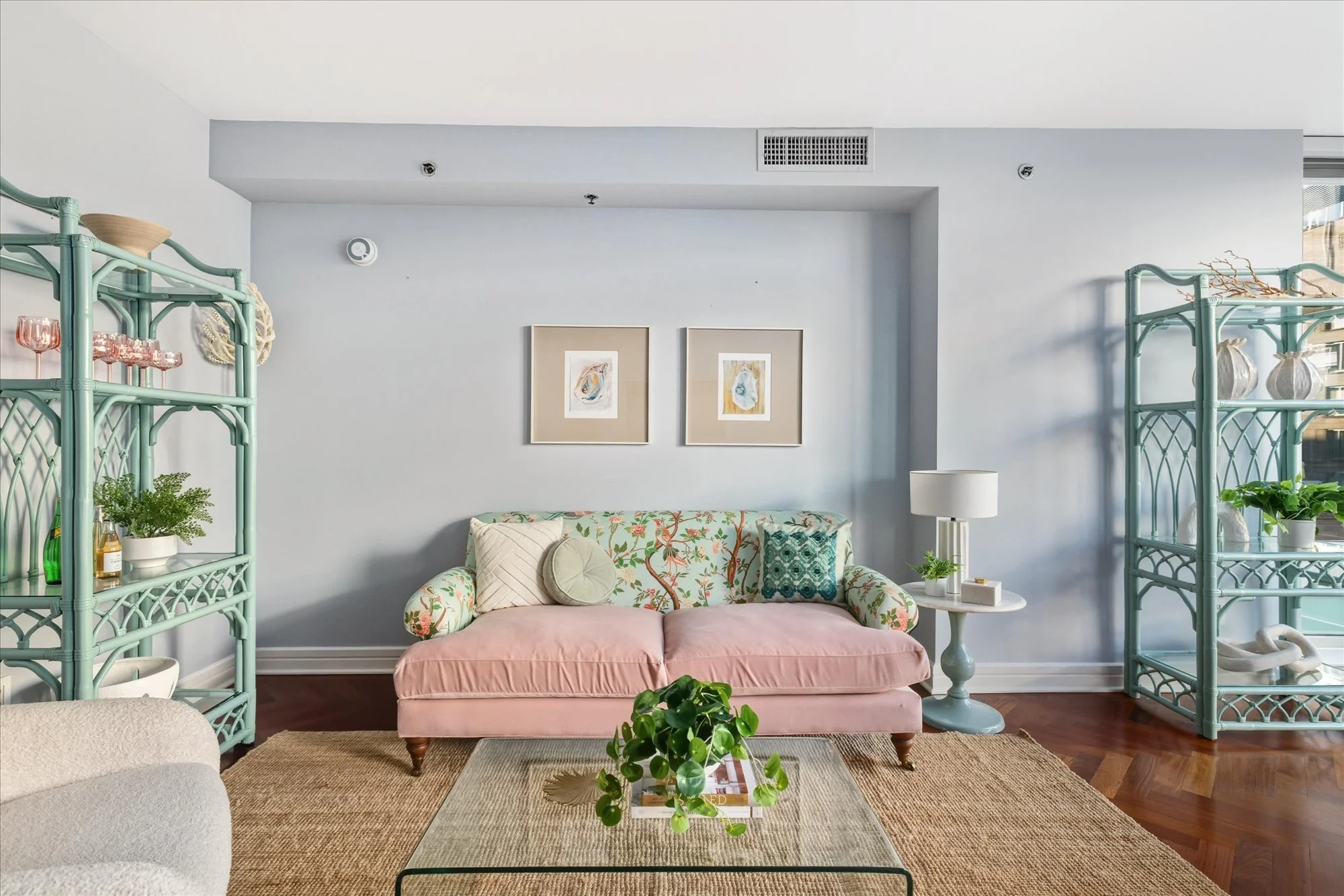 A living room with a pink floral couch and a beige armchair, a glass coffee table with a small green plant and magazines, light blue shelving units on both sides, white walls with two framed abstract art pieces, a white side table with a lamp, and wo