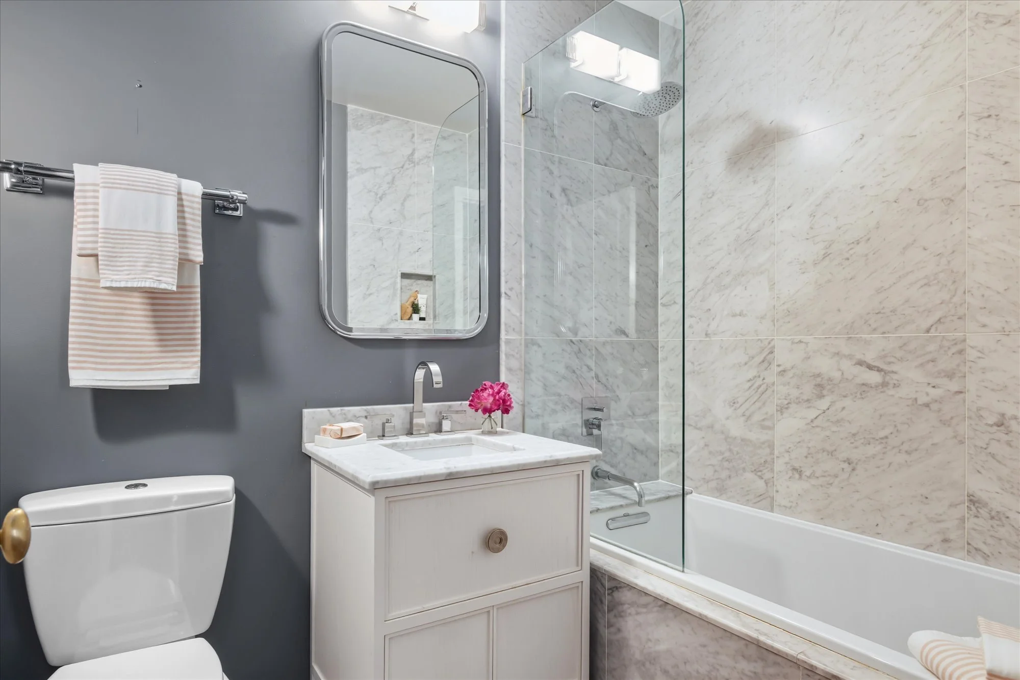 Modern bathroom with a gray wall, white vanity with marble countertop, pink flowers, large rectangular mirror, towel rack with striped towels, toilet, marble bathtub with glass shower partition, and marble tiles on the wall.