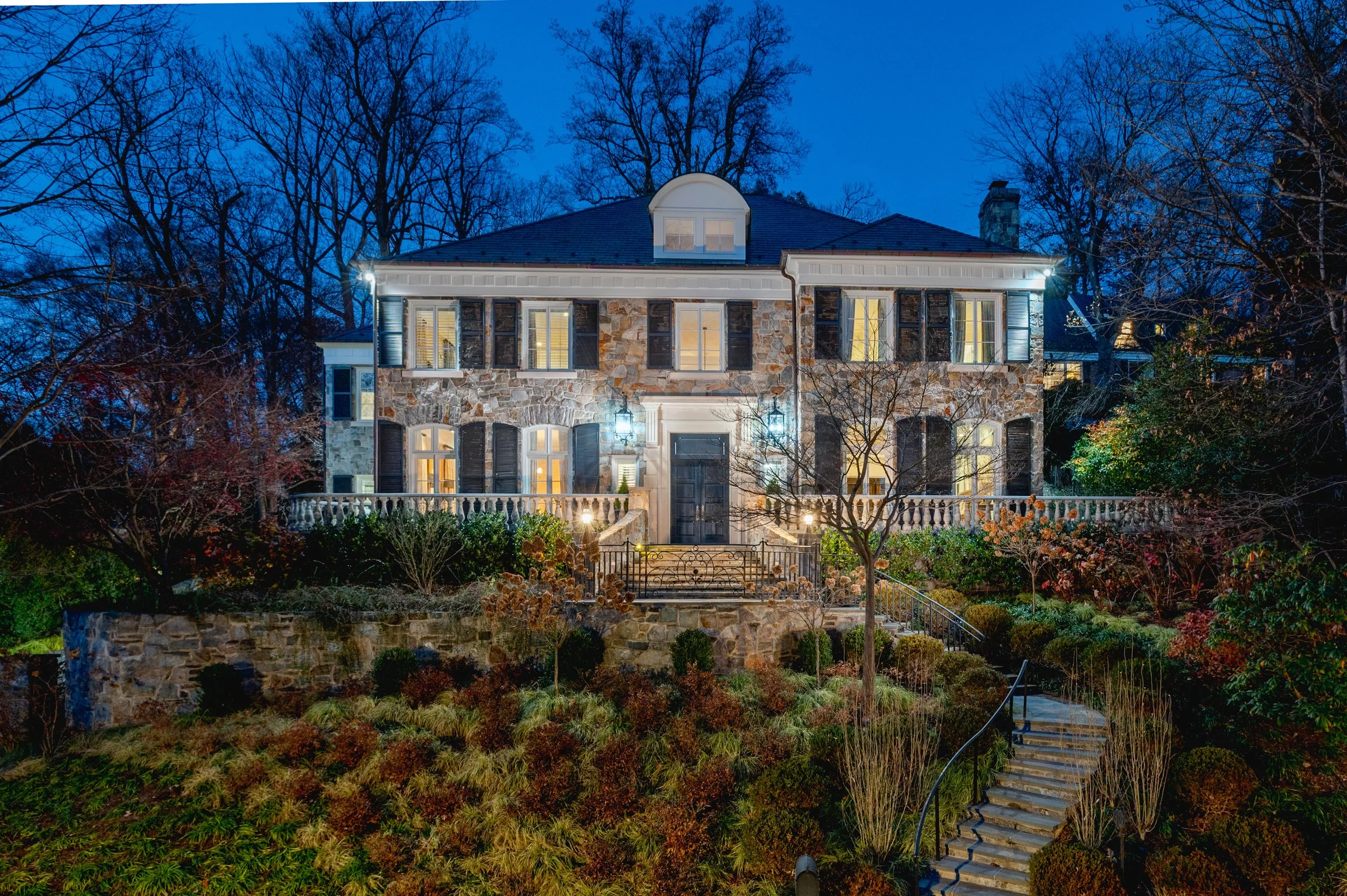 A two-story stone house with white trim and black shutters, illuminated from within, surrounded by trees, shrubs, and a garden at night with exterior lighting.