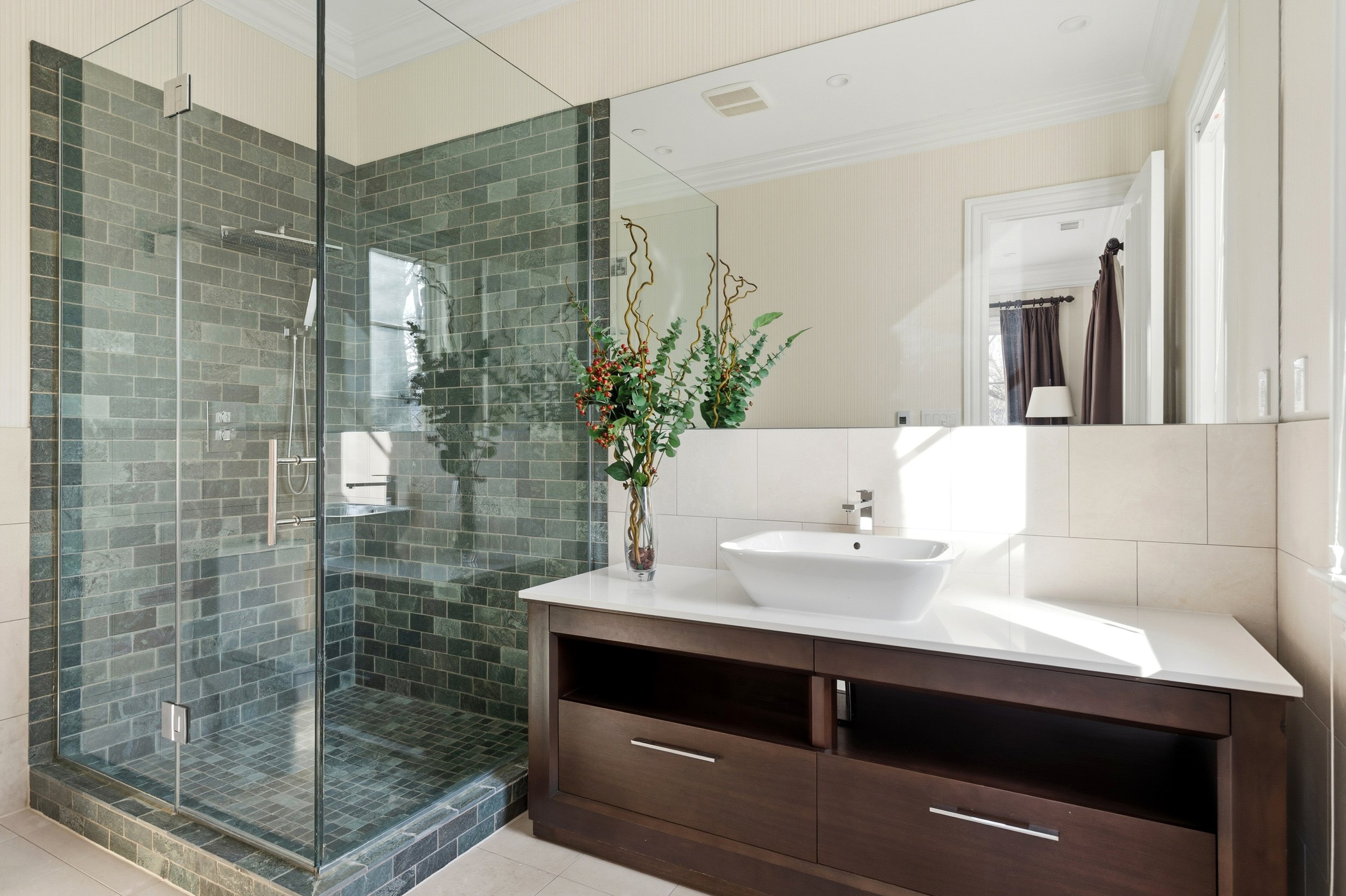 Modern bathroom with a walk-in shower featuring greenish-gray tiles and a glass door, a white vessel sink on a wooden vanity, a large mirror, a vase with greens and red berries, and a window with dark curtains.