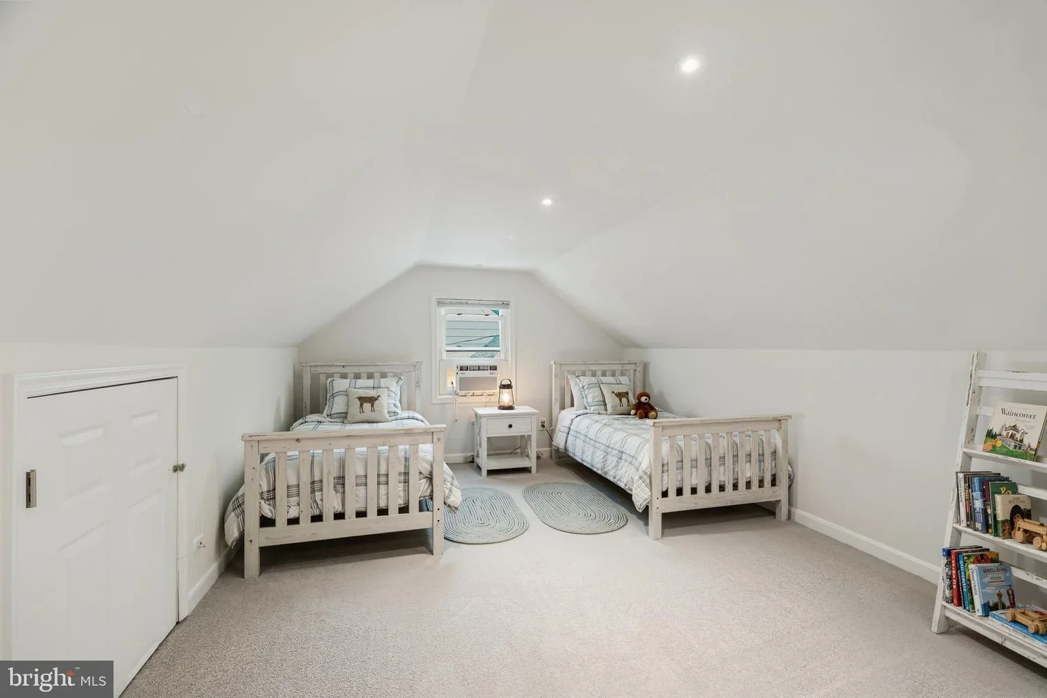 A cozy bedroom with two twin beds with white wooden frames, plaid bedding, and decorative pillows, a small white nightstand with a lantern, and a bookshelf with books, set in an attic with sloped ceiling and white walls.