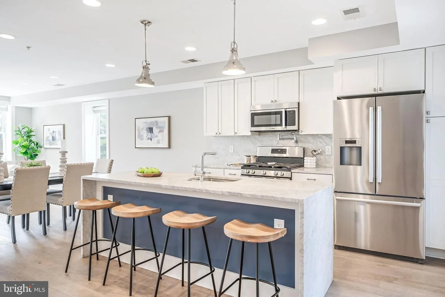 Modern kitchen with white cabinets, stainless steel refrigerator, microwave, and stove, marble countertops, island with three wooden bar stools, dining area with beige chairs, and artwork on the wall.