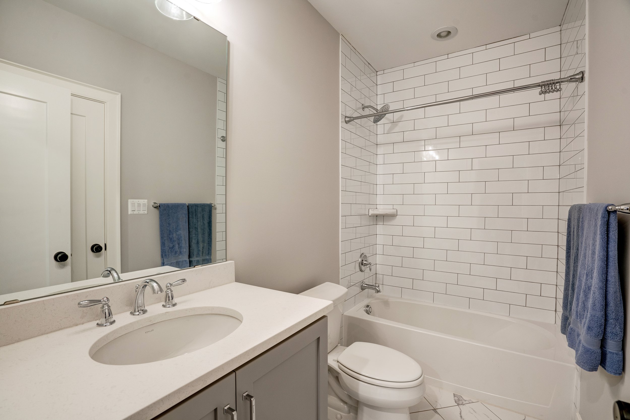 A bathroom with a white countertop, a sink, and a large mirror above it. There is a towel hanging on a bar on the wall. In the background, there is a bathtub with a showerhead and a curtain rod, with white subway tiles on the wall.