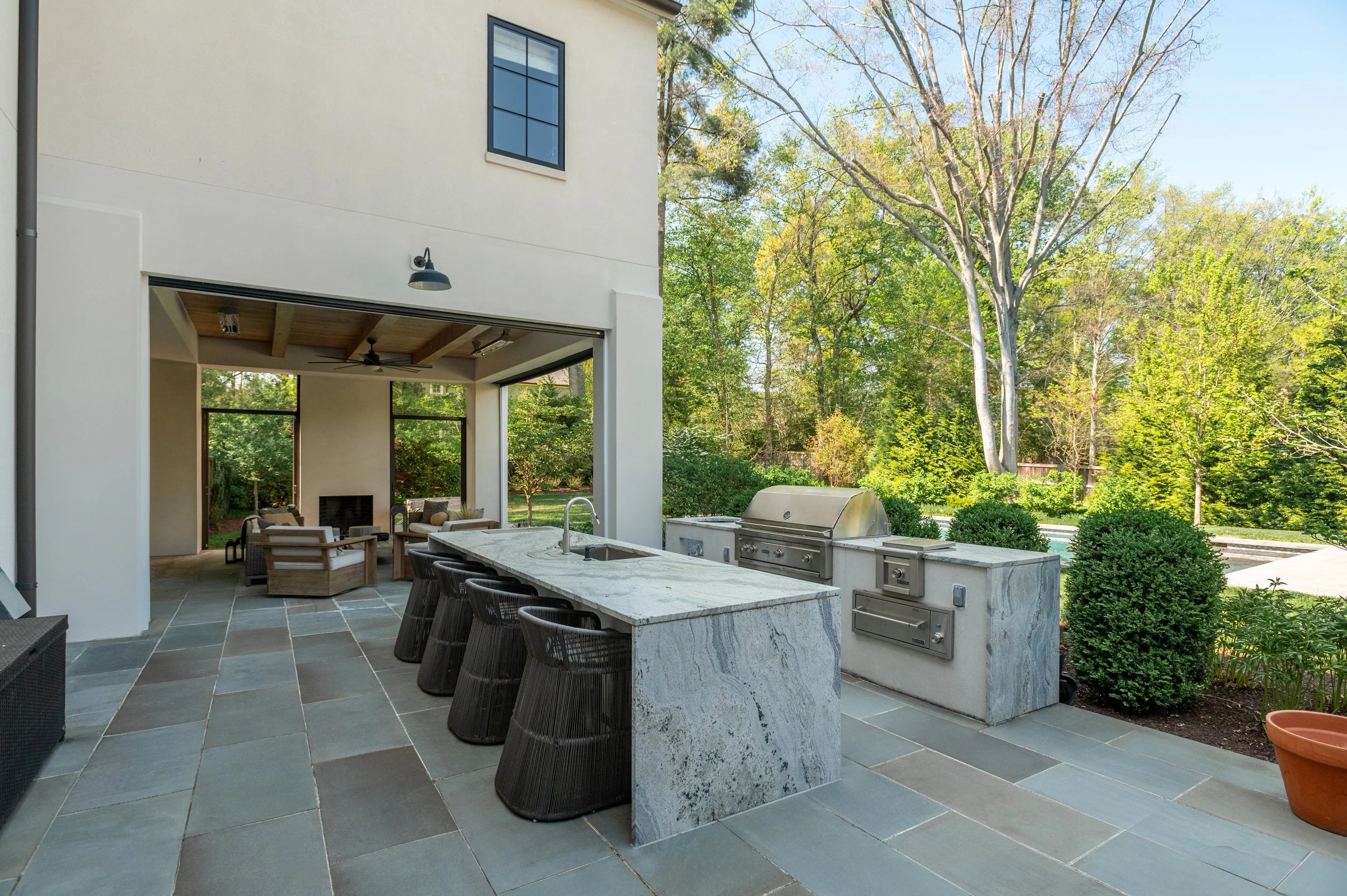 Outdoor patio with bar seating, kitchen grill, and seating area under a covered section, surrounded by trees and greenery.