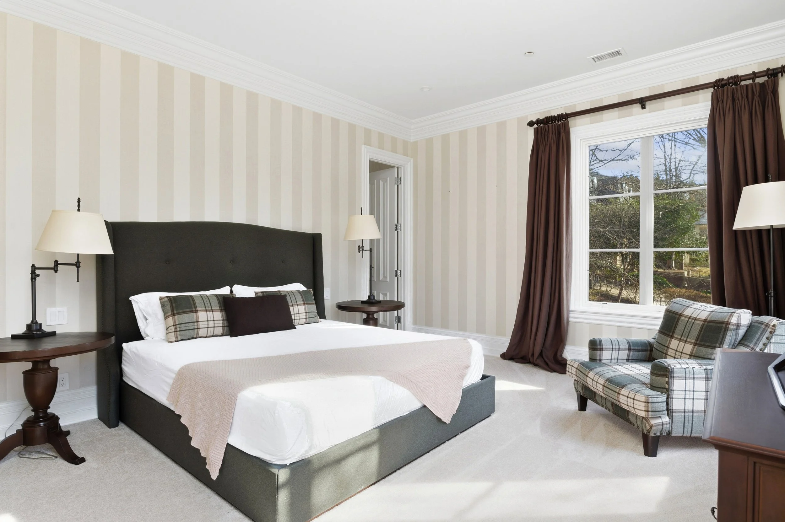 A bedroom with a large window with brown curtains, a bed with a dark upholstered headboard and plaid pillows, two matching bedside lamps, a plaid armchair, and a door in the corner.