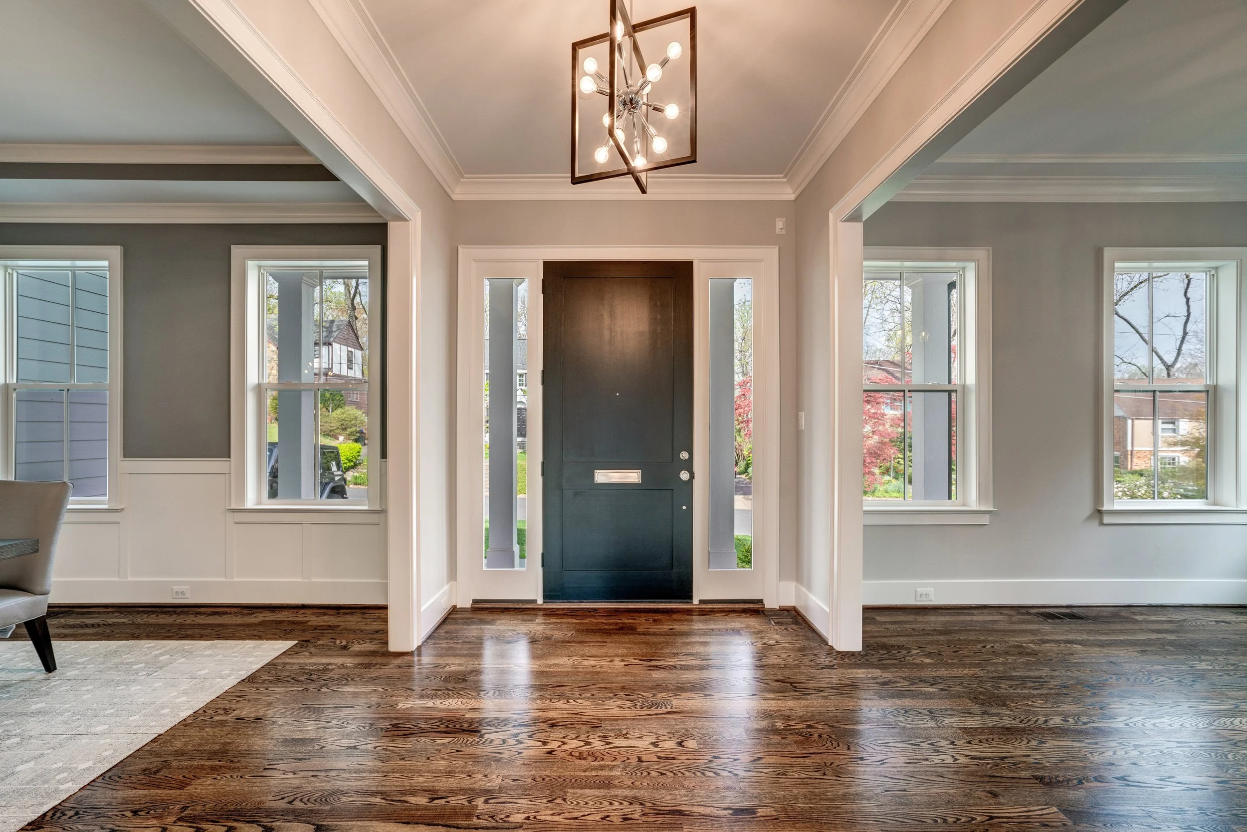 View of a modern home entryway with hardwood flooring, a black front door with a mail slot, and large windows on each side allowing natural light in. A geometric chandelier hangs from the ceiling, and outside trees can be seen through the windows.
