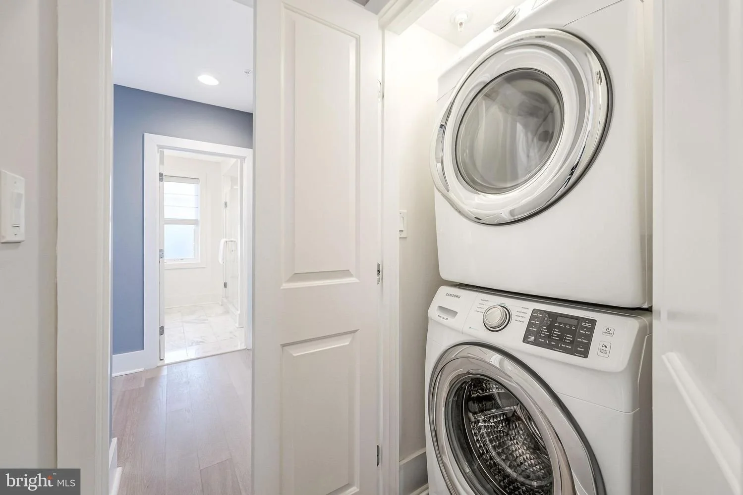Stacked white washing machine and dryer in a small laundry closet with doors open, adjacent to a hallway leading to a bathroom with a window, in a modern home.