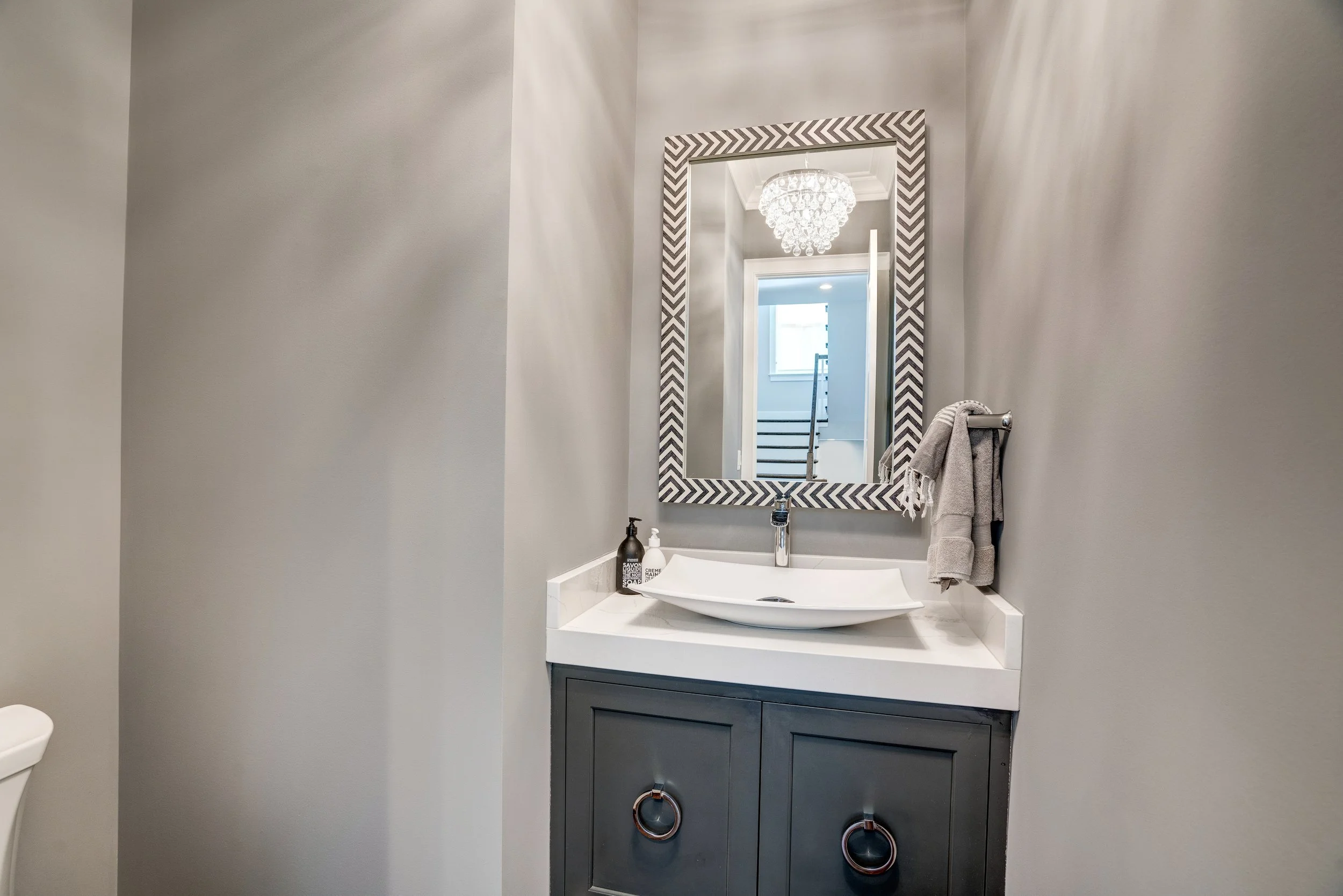 A modern bathroom vanity with a white sink, a dark gray cabinet with circular copper handles, and a large mirror with a chevron-patterned frame. The mirror reflects a chandelier and a staircase in the background.