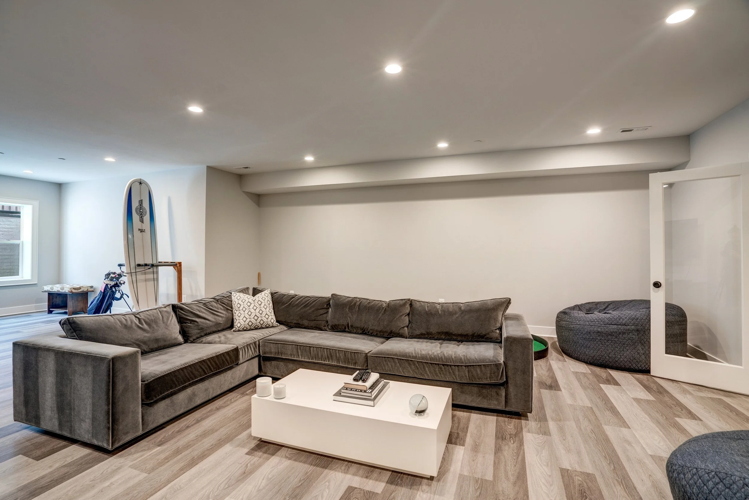 Living room with a large gray sectional sofa, a white coffee table, a bean bag, and a wall with various sports memorabilia, including a surfboard and a baseball bat, illuminated by ceiling lights.