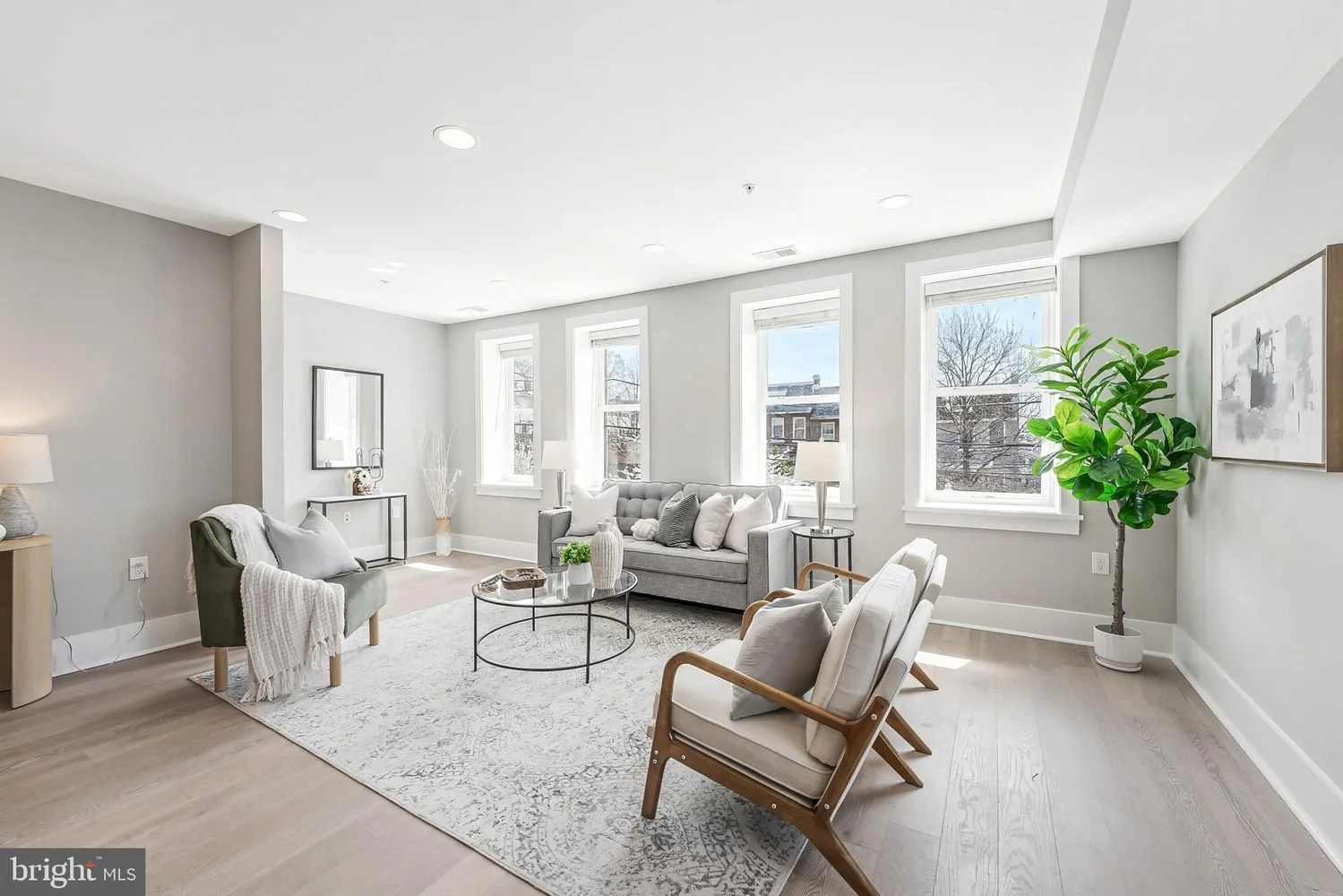 Bright living room with white walls, hardwood floors, large windows, gray sofa, white armchairs with wooden arms, round glass coffee table, potted plant, and modern artwork