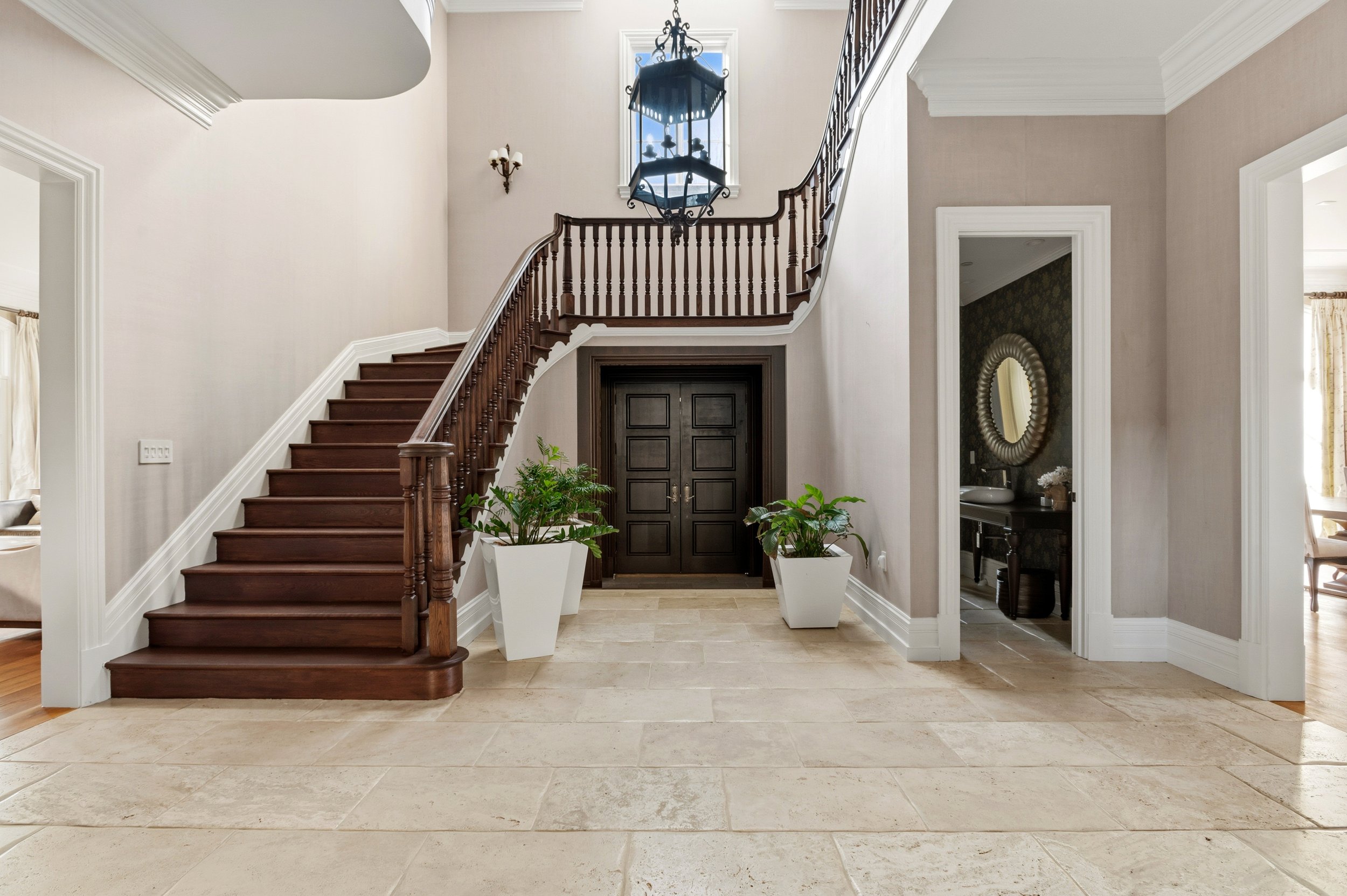 Interior view of a home entryway with a curved staircase, dark brown wooden steps, white trim, and a black chandelier. Two large white planters with green plants are at the bottom of the stairs, and a dark double door is visible beneath the staircase