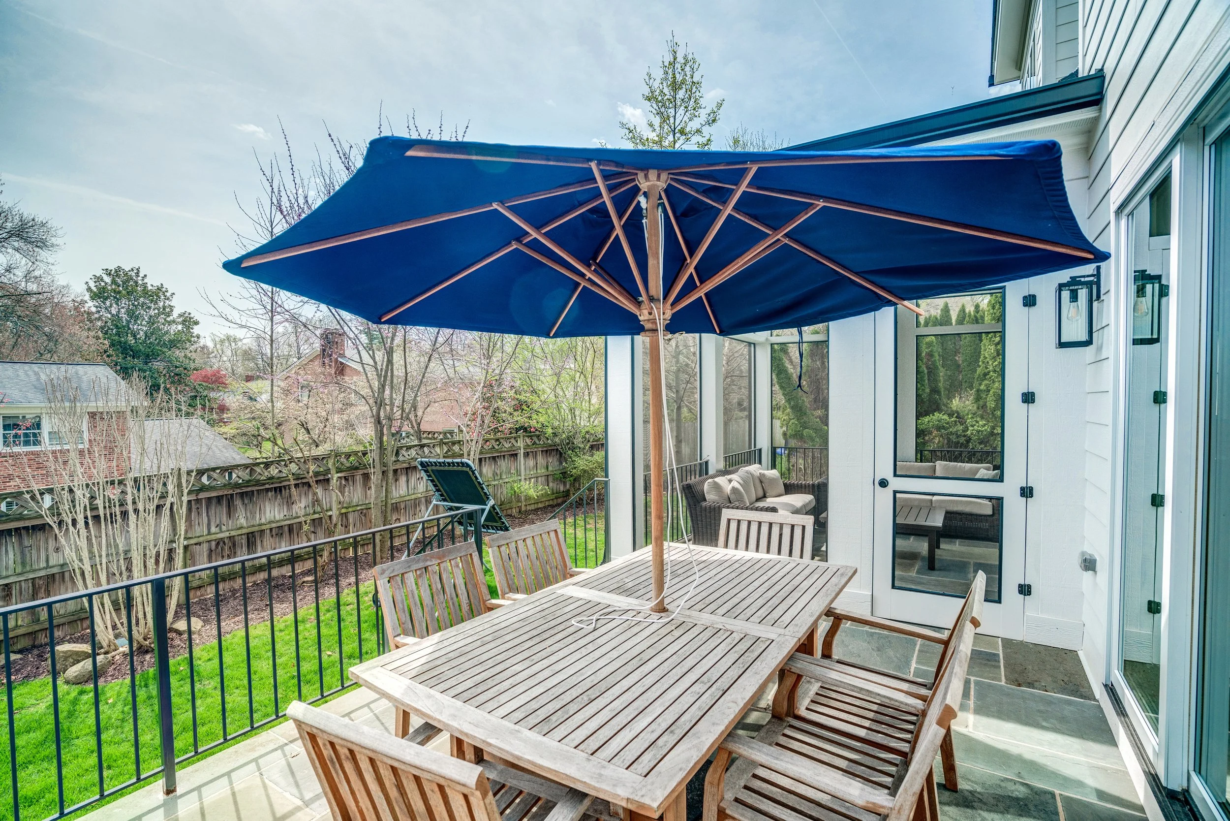 Outdoor patio with wooden table and chairs, blue patio umbrella, surrounded by a backyard fence, trees, and neighboring houses, adjacent to a house with white siding and a sliding glass door.