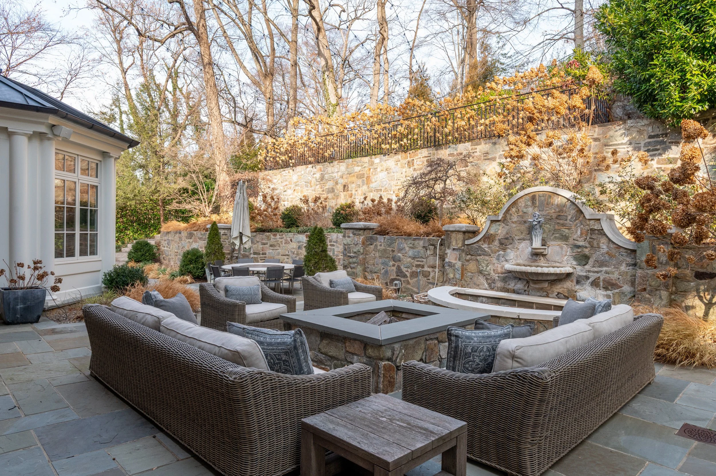 A backyard patio with outdoor furniture, including a sectional sofa, chairs, and a fire pit, surrounded by stone walls and a garden with trees and shrubs.