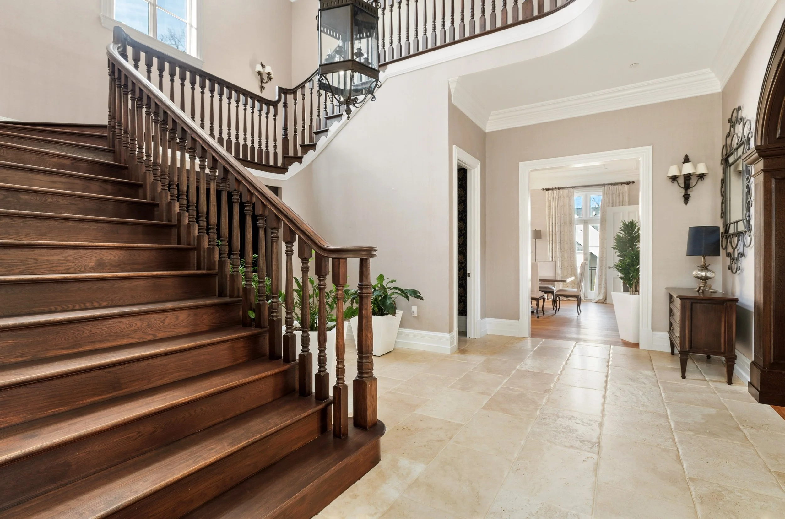 Interior view of a house entryway with a wooden staircase, beige tiled floor, and a view into a dining area with windows and curtains.