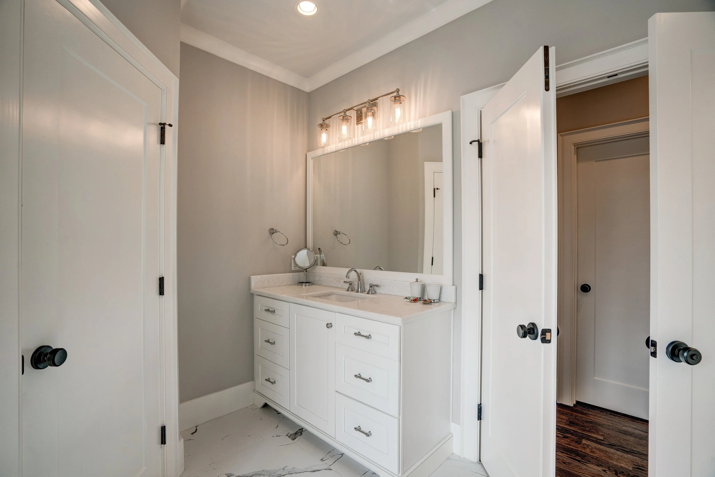 Modern bathroom with a large mirror, vanity with sink, and light fixtures above the mirror. The walls are painted light gray, and the floor has white marble tiles with gray veining. There are two partially open white doors with black hardware.