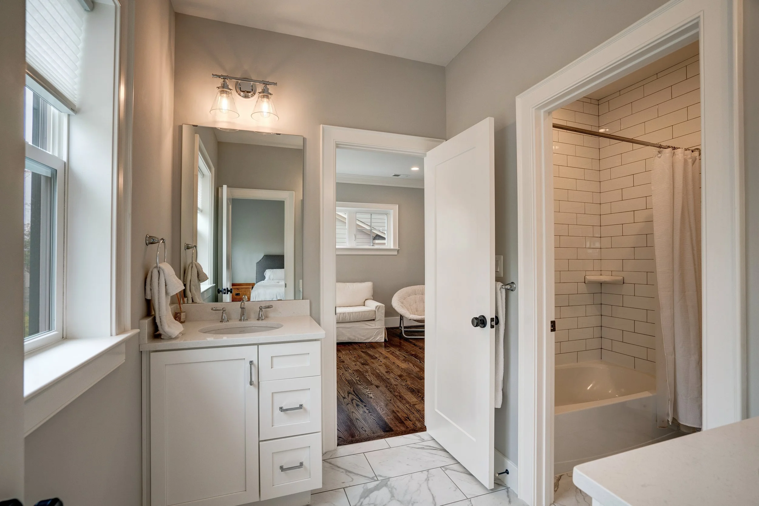 Bathroom with a white vanity, two mirrors, towel hooks, a shower with a curtain, a window, and a view of a bedroom with a bed and a chair inside.