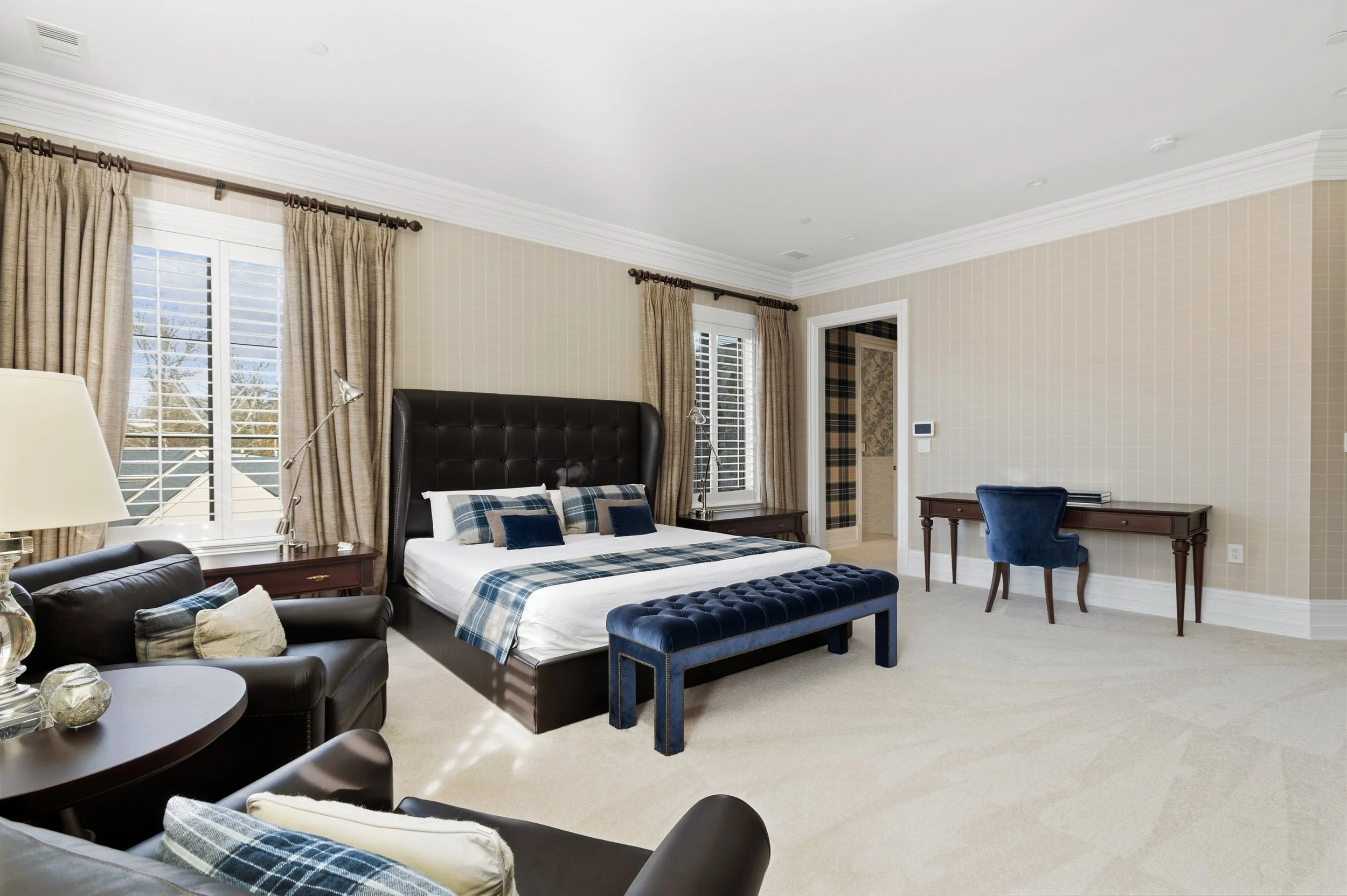 A bedroom with a large bed, dark headboard, plaid bedding, nightstands, beige curtains, and windows with white shutters; also includes a desk with a chair, a small sitting area with leather chairs, and decorative items.