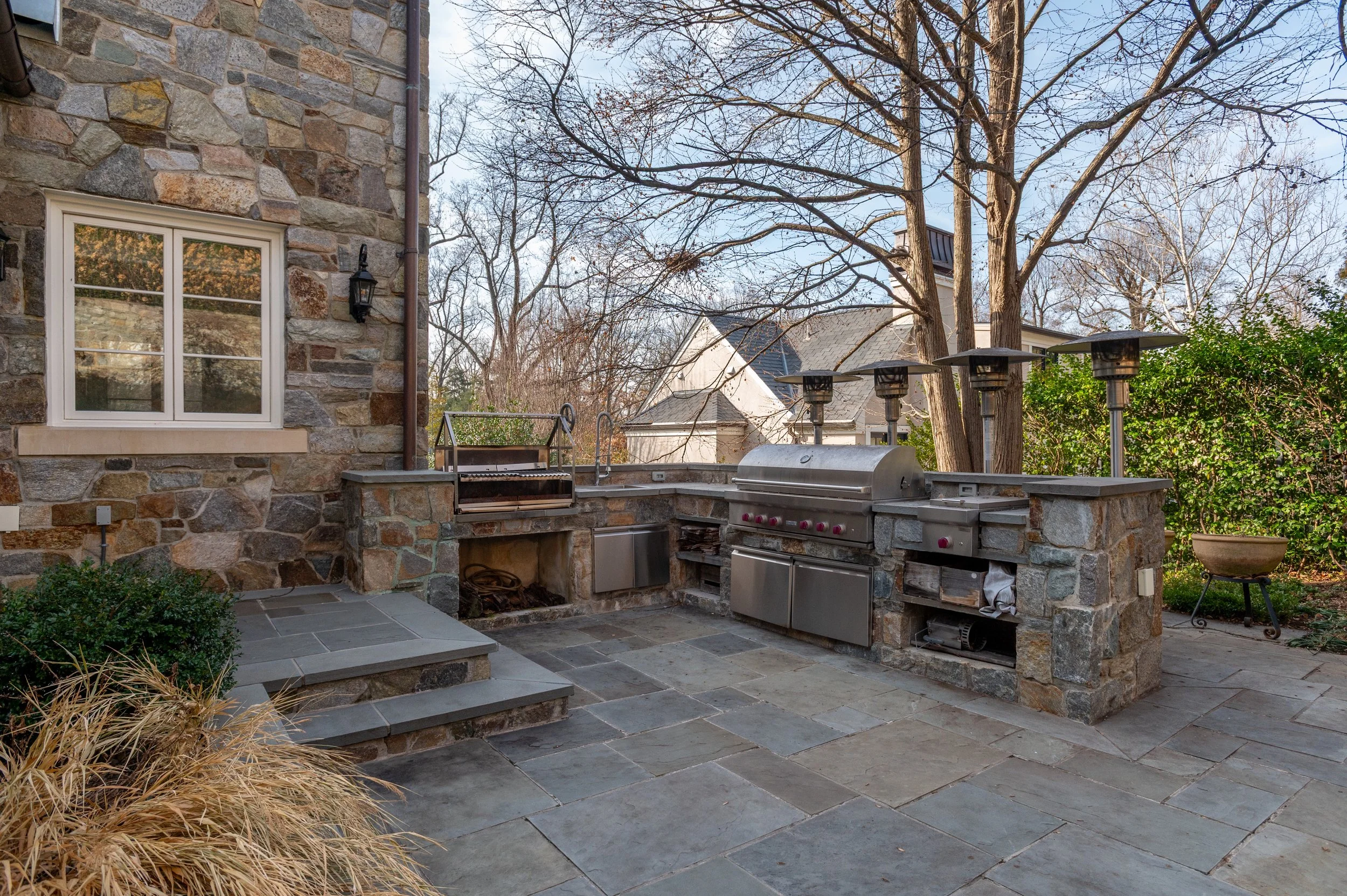 A backyard patio area with stone flooring, a built-in stone outdoor kitchen, a grill, and four patio heaters. There is a stone house wall with a window and a lamp, a tree with bare branches, and some greenery and plants around.