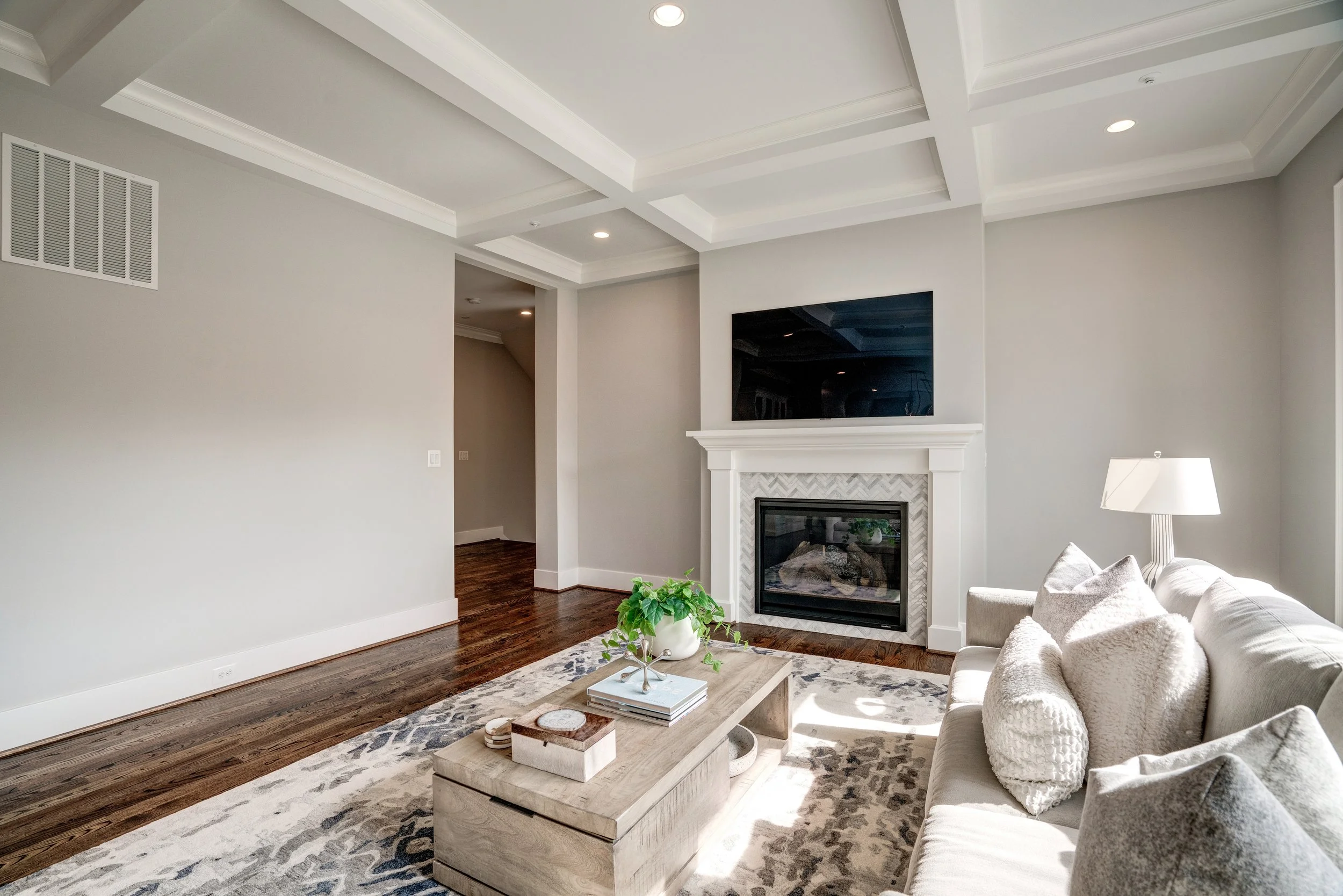 Living room with white walls, a fireplace with a TV mounted above, a white sofa with pillows, a coffee table with decor, a lamp, a rug, and hardwood floors.