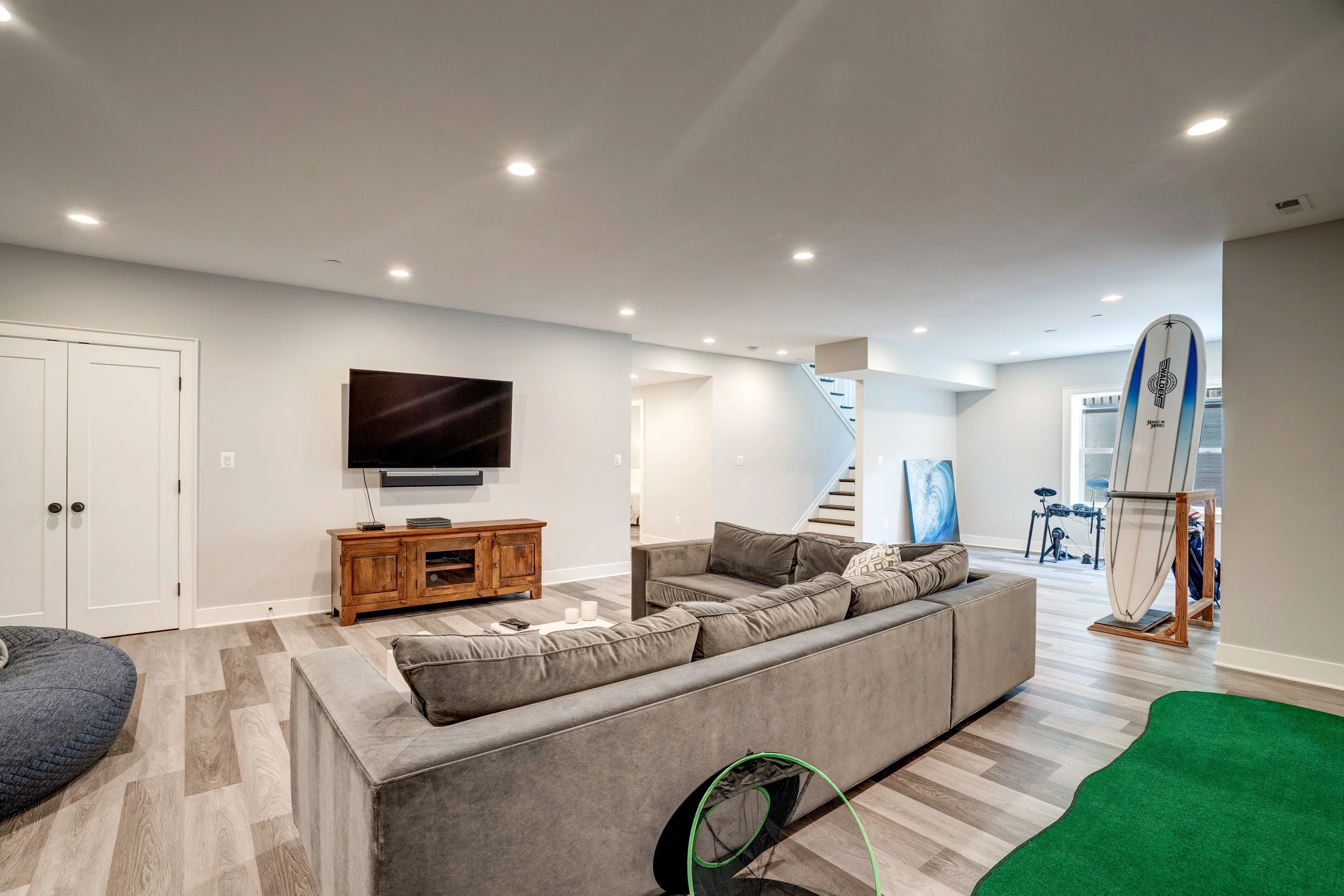 Living room with gray couches, a wooden TV stand, a flat-screen TV, and a surfboard in the corner, with a staircase and window in the background.
