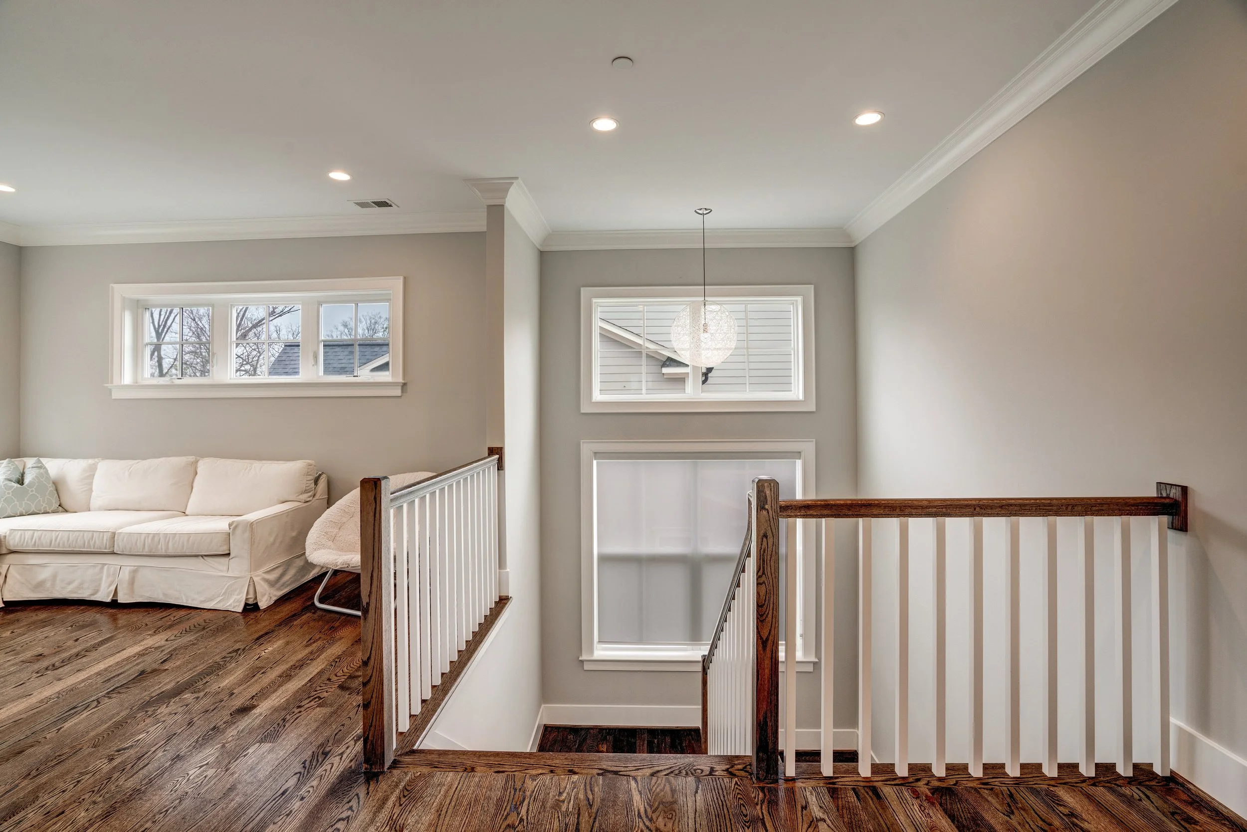 Interior of a house with a staircase, windows, a white couch, and a hanging light; hardwood floors and light-colored walls.