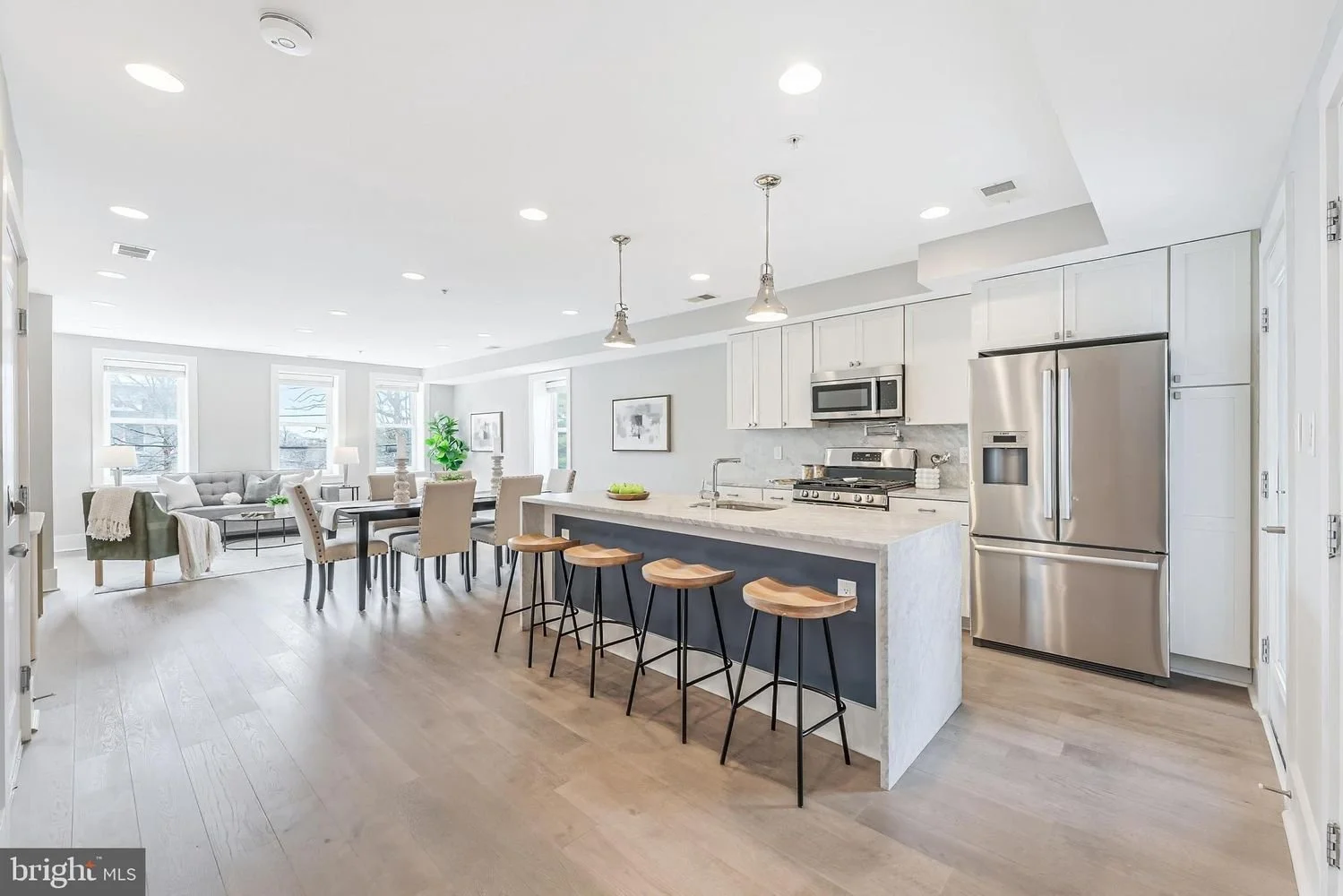 Open-concept kitchen and living room with white cabinetry, stainless steel appliances, marble island, beige bar stools, dining table, and a gray sofa in a bright space with large windows and light wood flooring.