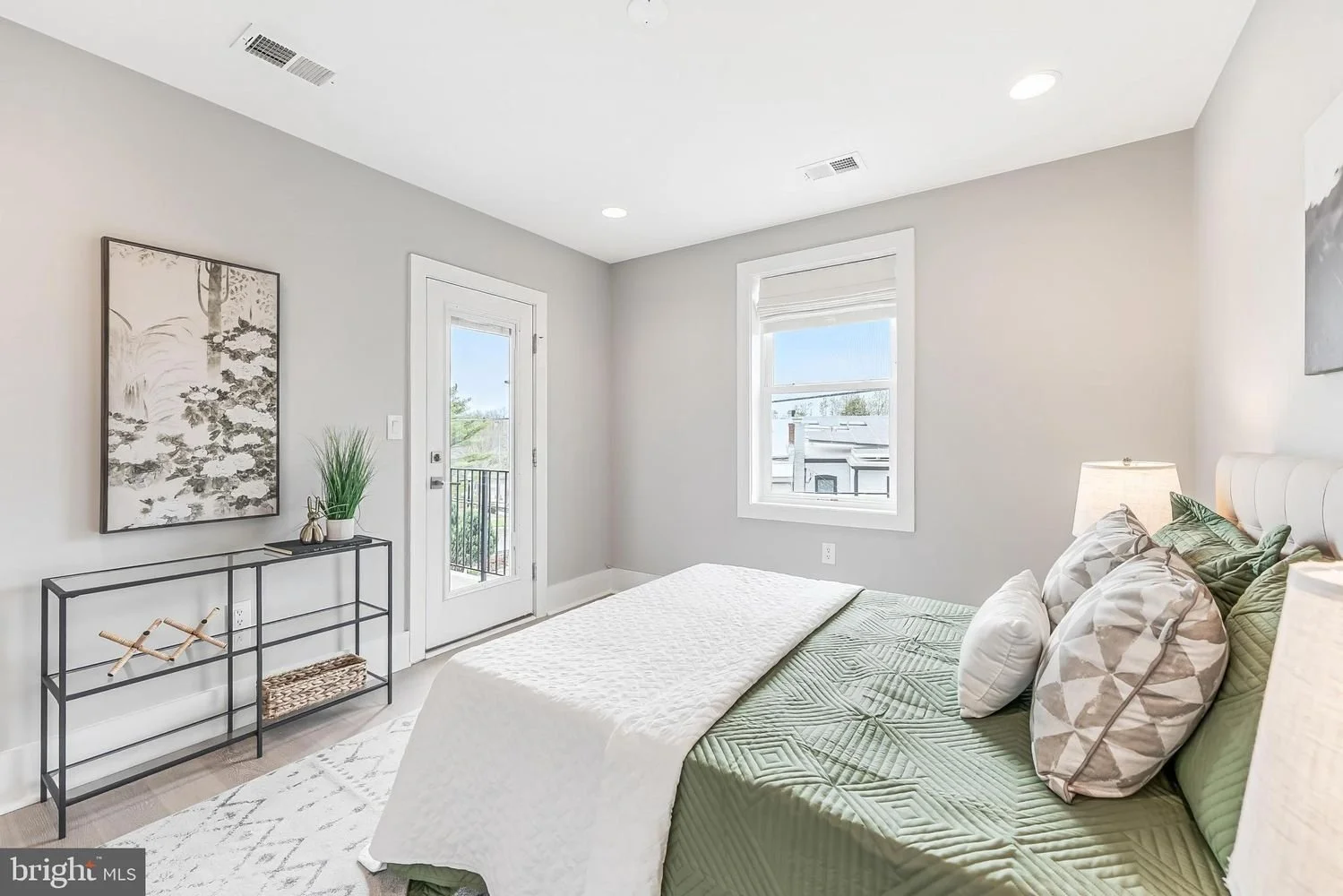 Bright bedroom with a bed, pillows, a nightstand with a lamp, a wall art, a potted plant on a metal shelf, and a door leading to a balcony with a view outside.