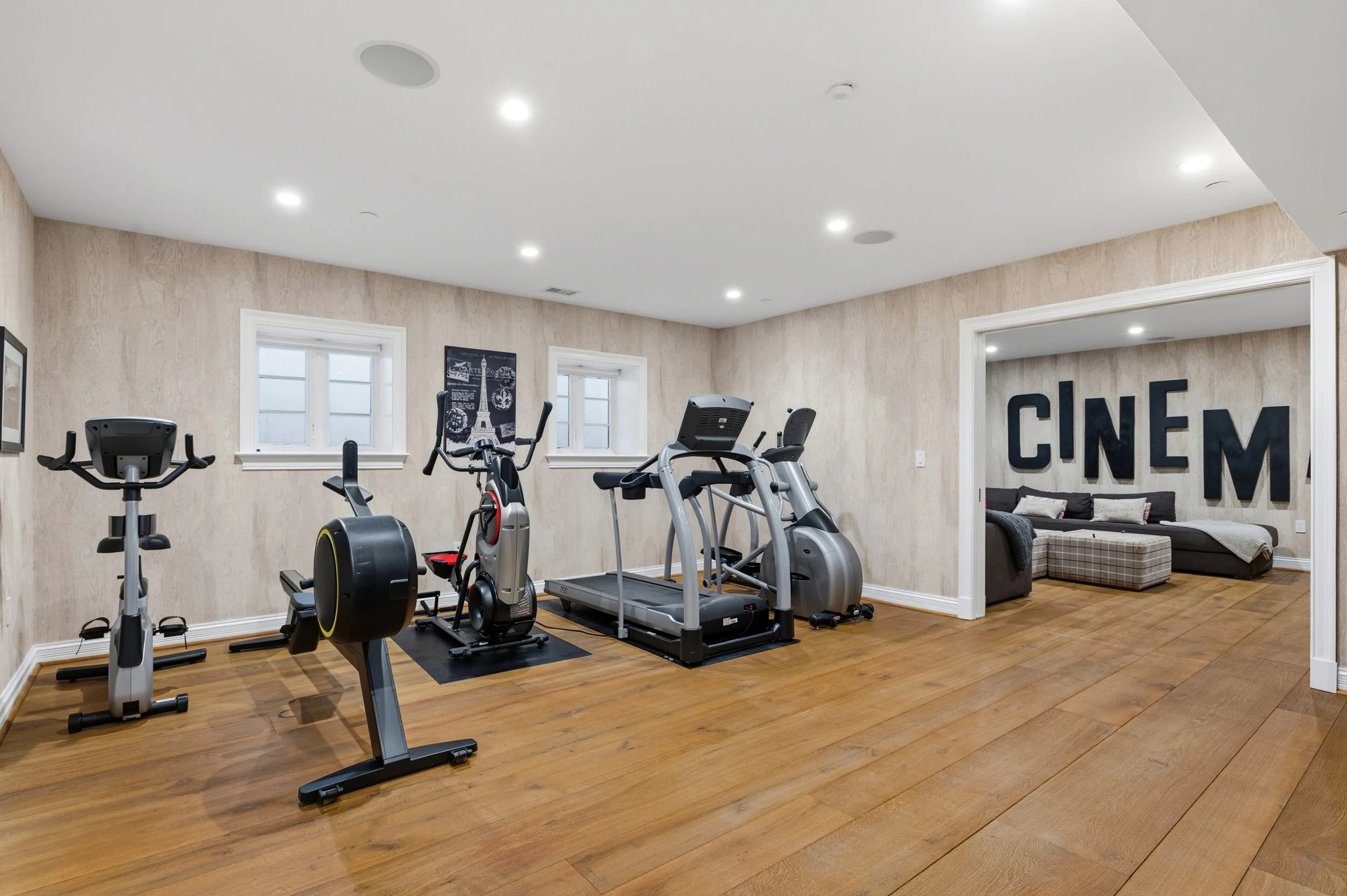 Home gym room with exercise equipment including a treadmill, elliptical, and stationary bike on wooden floors, beige walls with small windows, and a seating area with a sofa and large decorative letters spelling 'CINEMA' on the wall.
