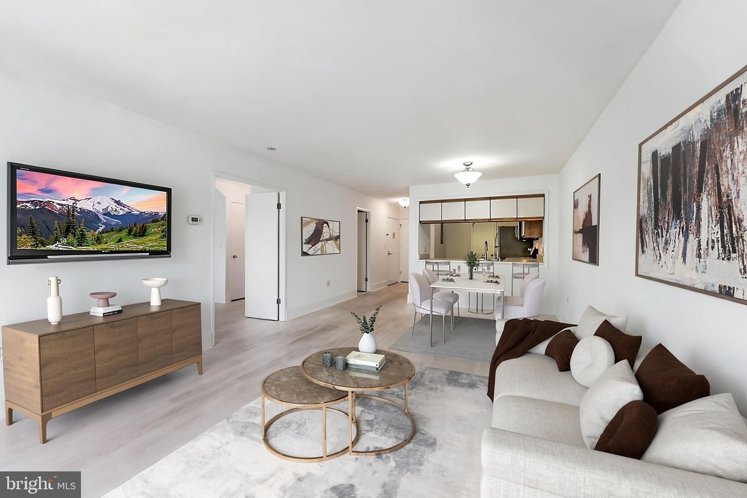 Bright and spacious living room with a wall-mounted TV, a beige sofa with brown and beige pillows, round nested coffee tables with decorative items, wall art, and an open plan kitchen and dining area.