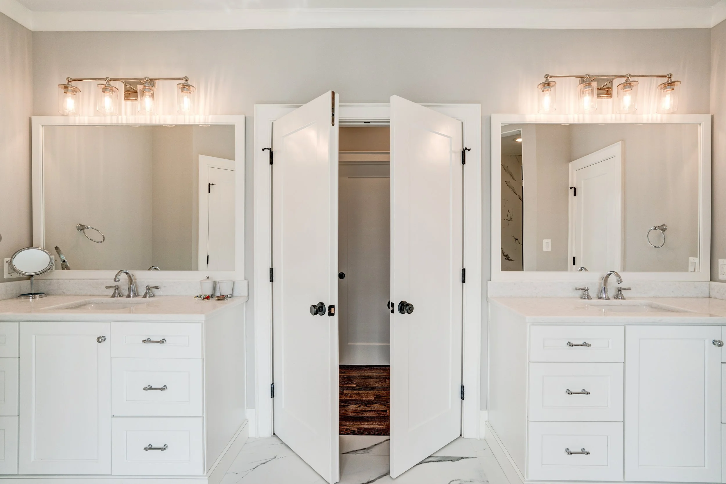 A bathroom with two white vanities, each with a mirror and light fixtures above, and a central open closet with doors. The: bathroom has marble countertops and flooring, and the closet reveals a dark wooden floor inside.