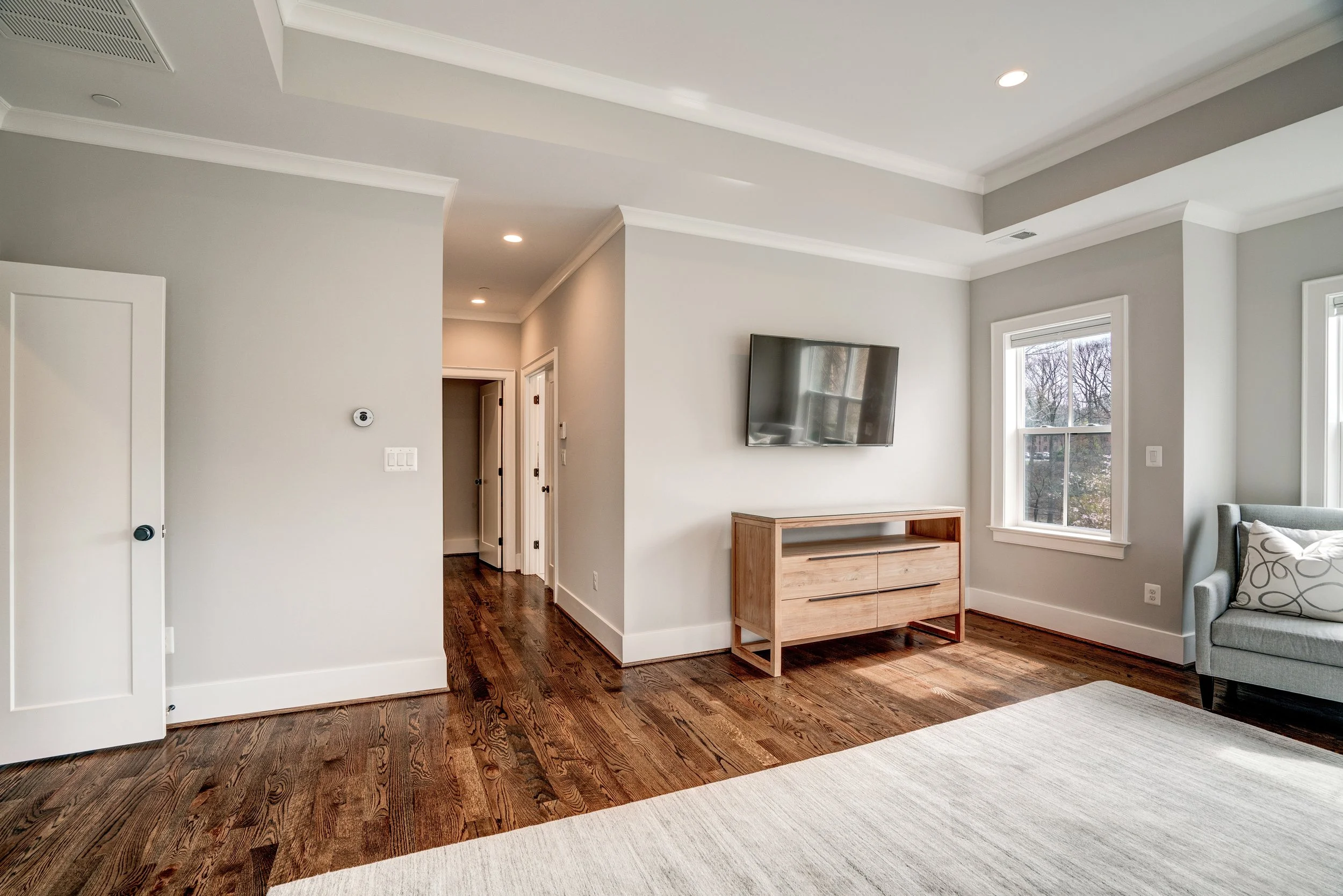 Living room with white walls, hardwood floors, a wall-mounted TV, a wooden TV stand, and large windows showing trees outside.