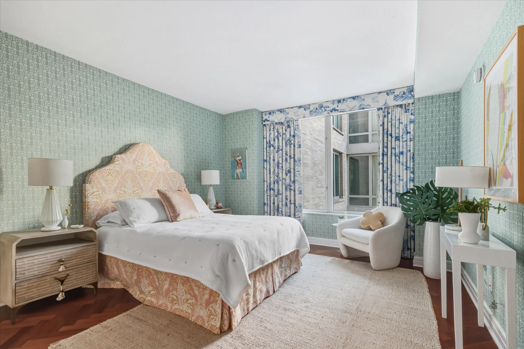 A bedroom with a bed that has a floral headboard, white bedding, and pillows. There are two white lamps on nightstands, a painting on the wall, and a window with blue and white curtains. A white armchair with a plush toy, a small table with potted pl