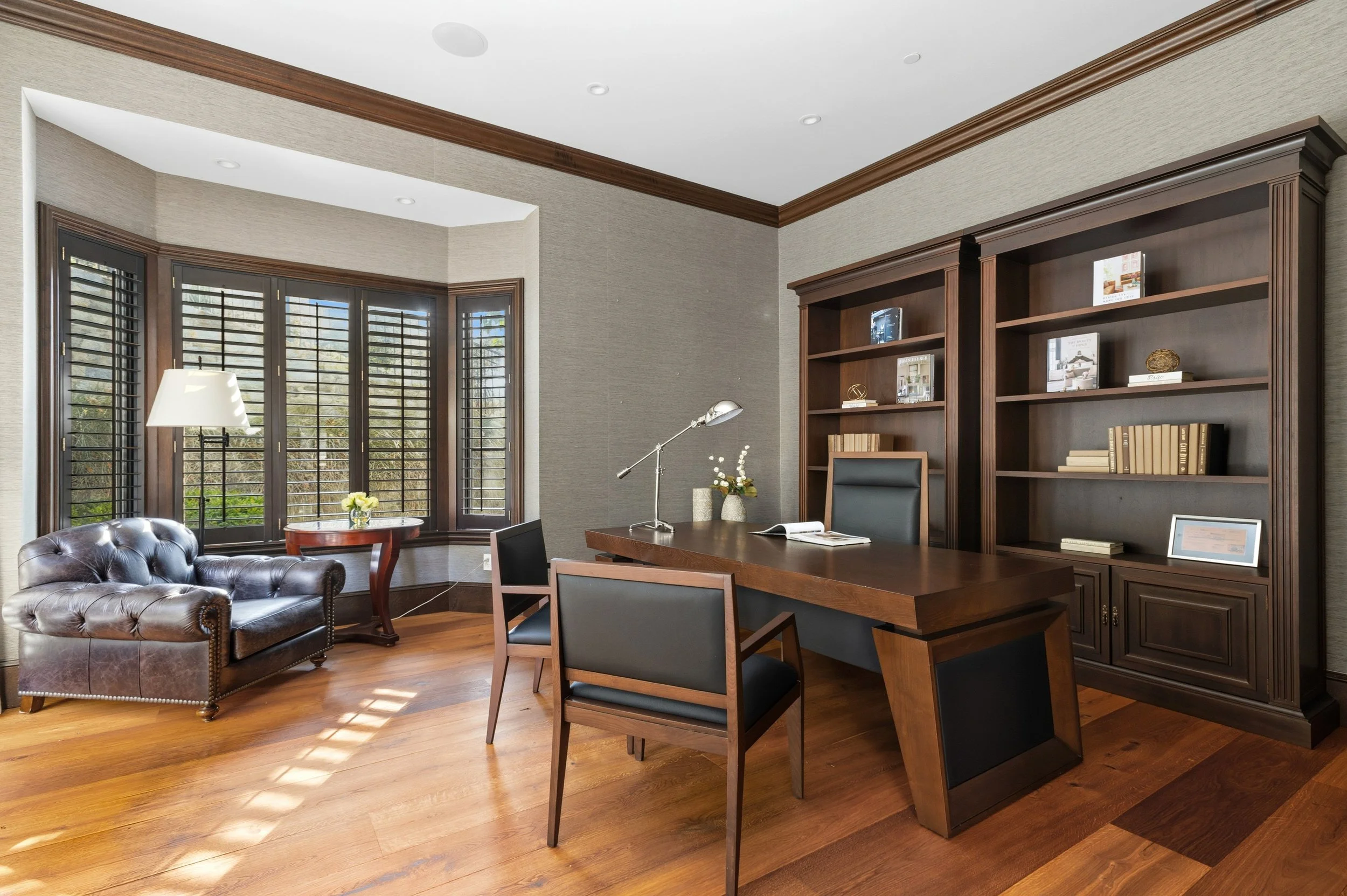 Office space with wooden flooring, large window with shutters, a leather tufted armchair, a round side table with flowers, a desk with chairs, a metal desk lamp, and a bookshelf.
