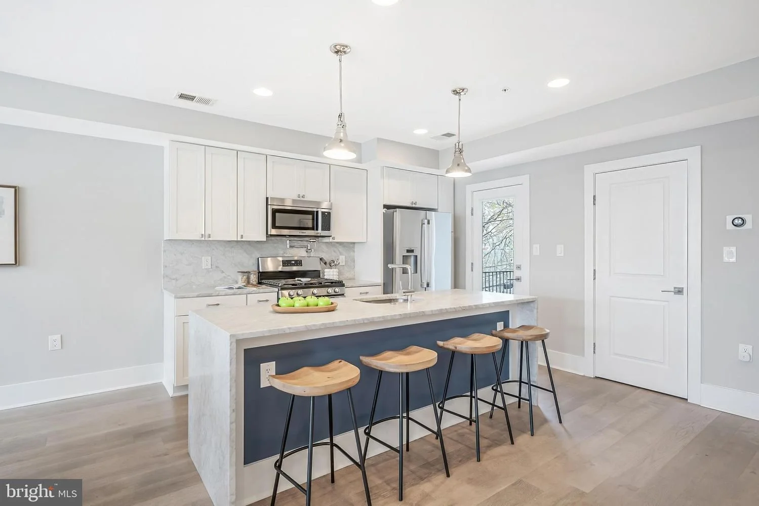 Modern kitchen with white cabinets, stainless steel appliances, gray walls, a kitchen island with a dark blue base, four wooden barstools, and a door leading outside.