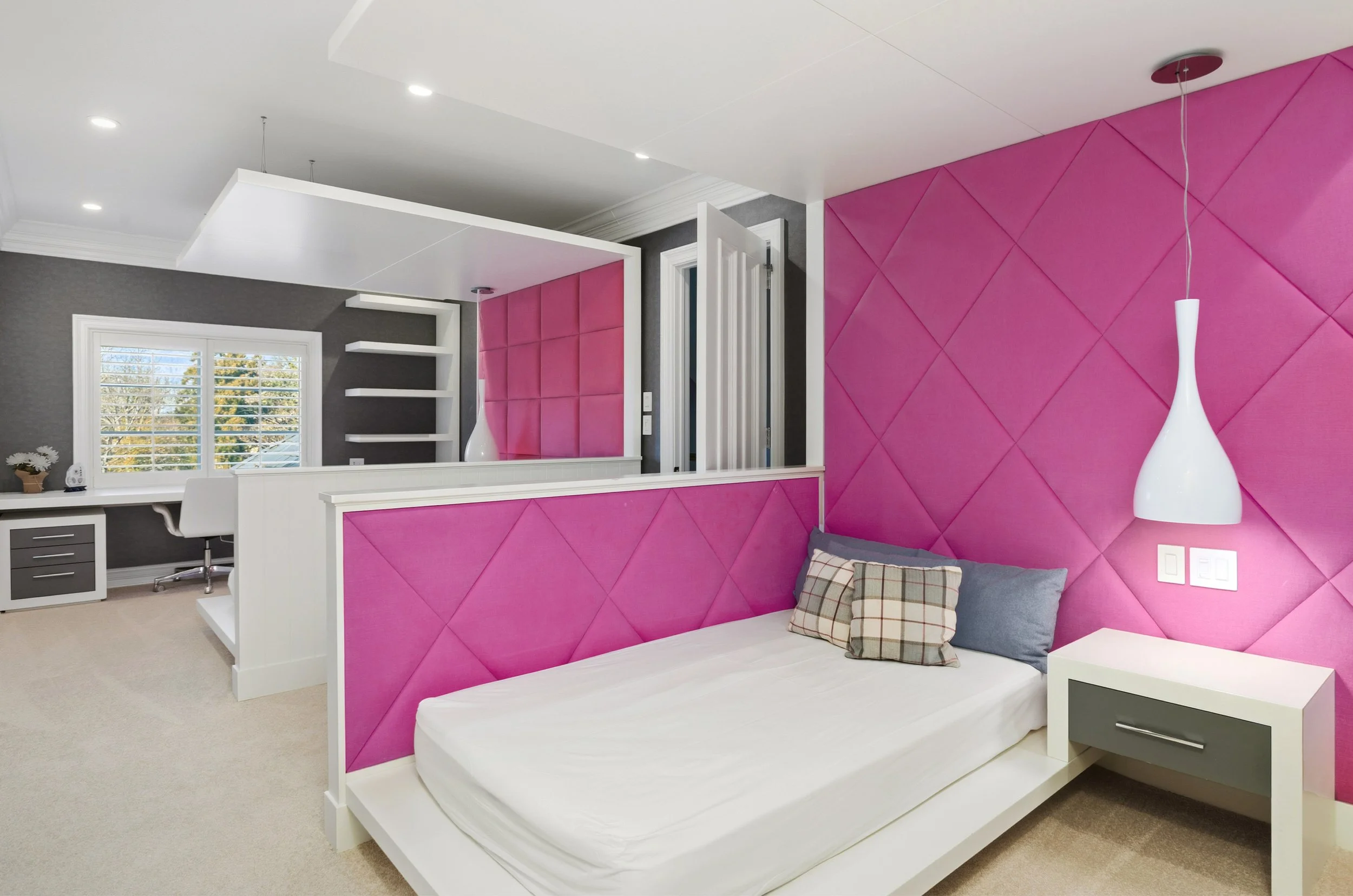 A modern bedroom with a pink, quilted accent wall, a white bed with a plaid pillow, a white hanging lamp, and a white office desk near a window showing trees outside.