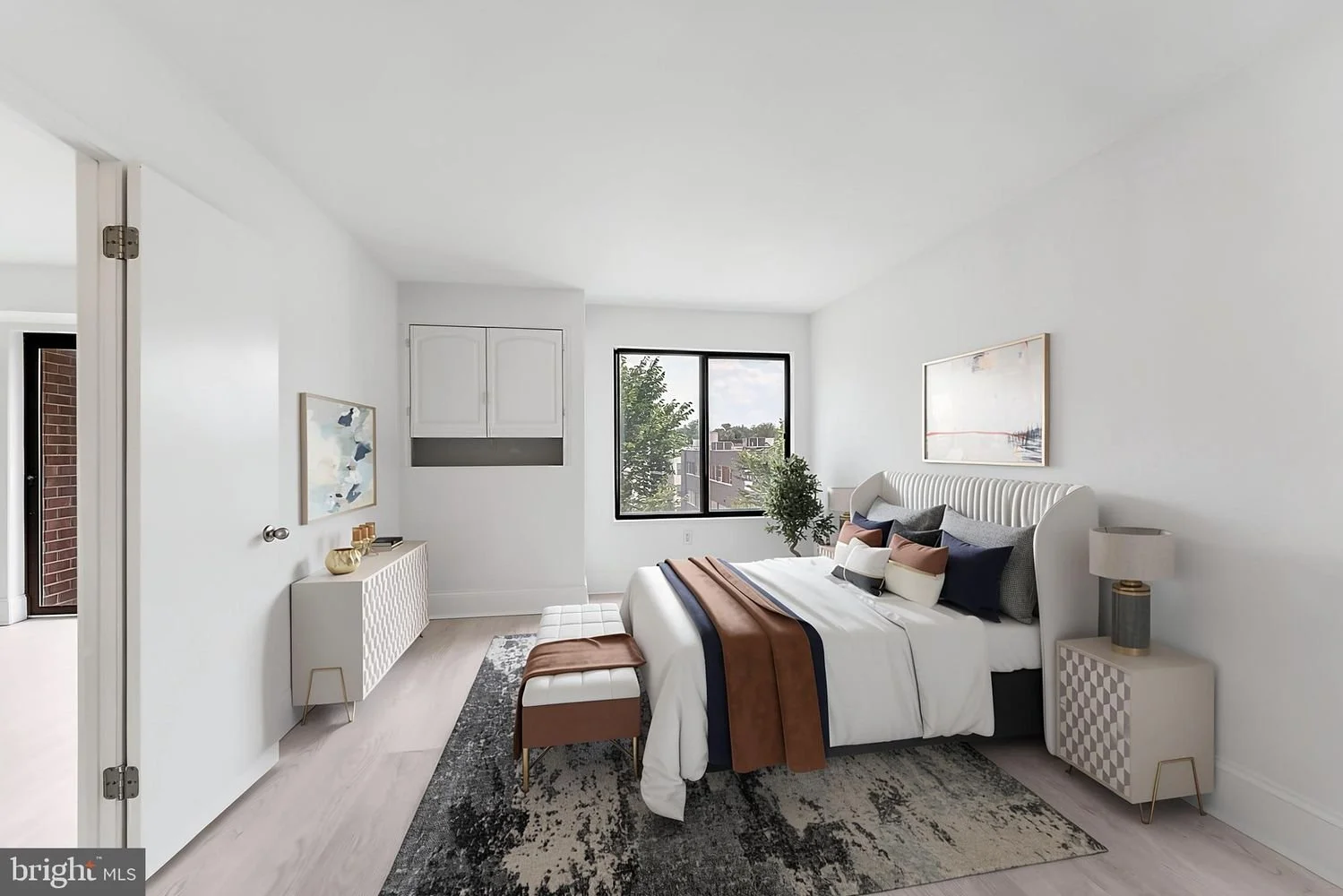 A modern bedroom with white walls, a bed with white, navy, and beige bedding, a bench at the foot of the bed, a nightstand with a lamp, and a large window showing trees and buildings outside.