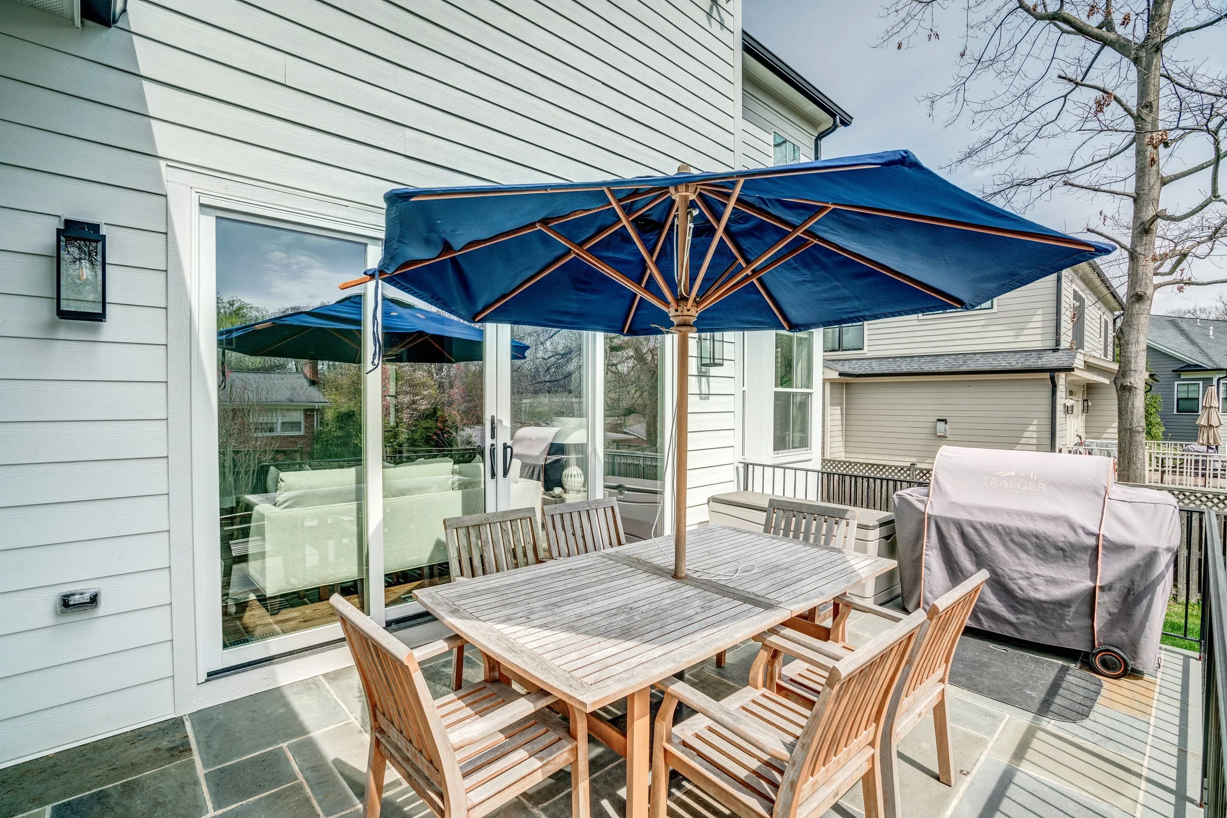 A wooden outdoor dining table with six matching chairs on a patio, shaded by a large blue patio umbrella, with house siding and trees in the background.