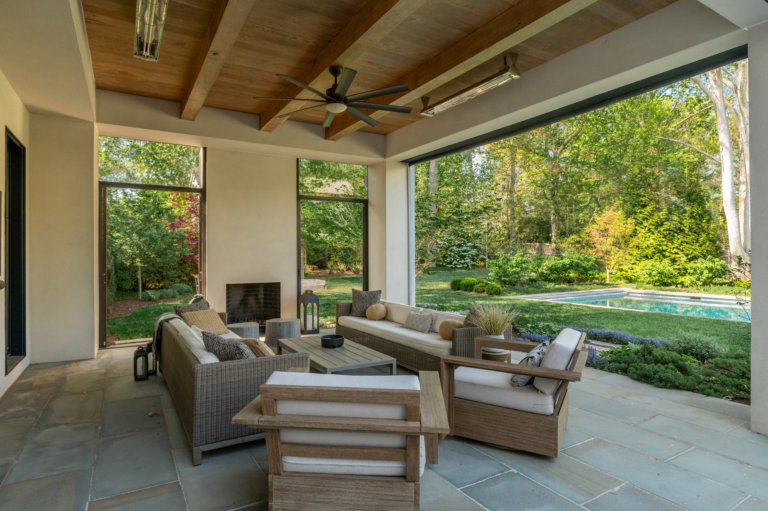 Covered outdoor patio with beige wicker seating, wooden accents, and large open windows looking out onto a lush green backyard with trees and a swimming pool.