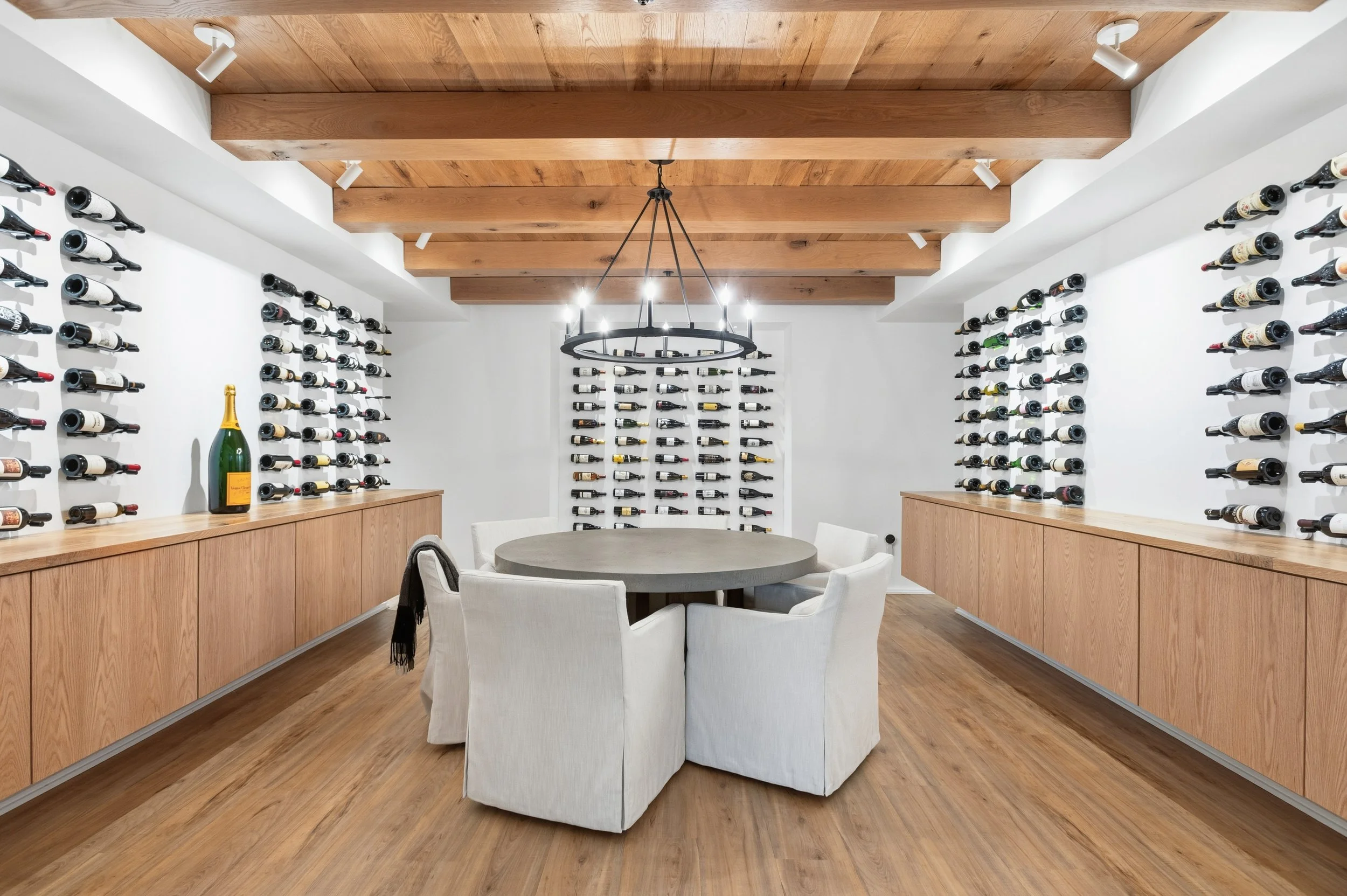 A wine cellar with white walls, wooden beams on the ceiling, and wall-mounted wine racks filled with bottles. A round table surrounded by white chairs is in the center, and a large champagne bottle is on the wooden sideboard.