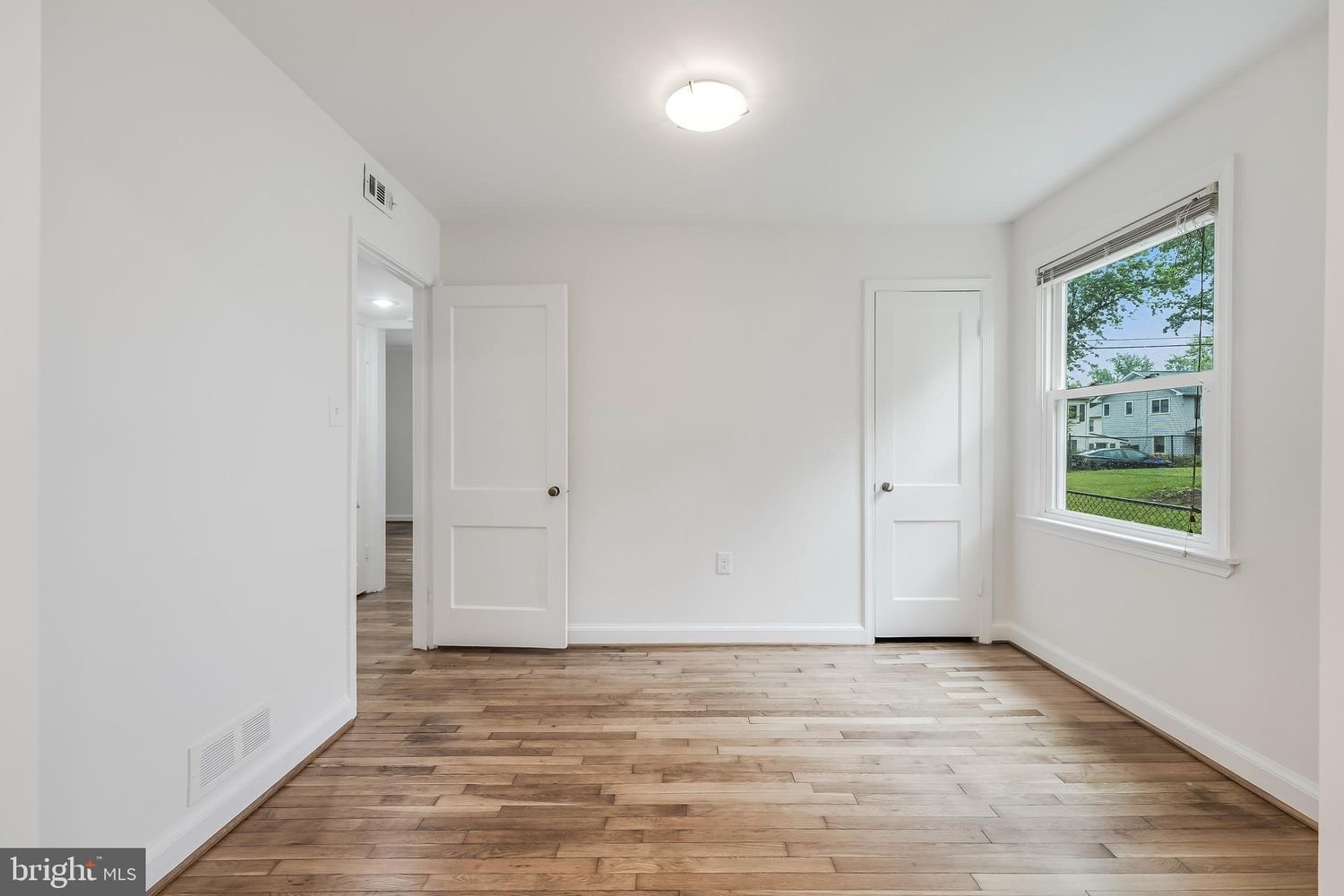 Empty room with hardwood floors, white walls, two closed doors, and a large window showing a grassy yard and neighboring houses.