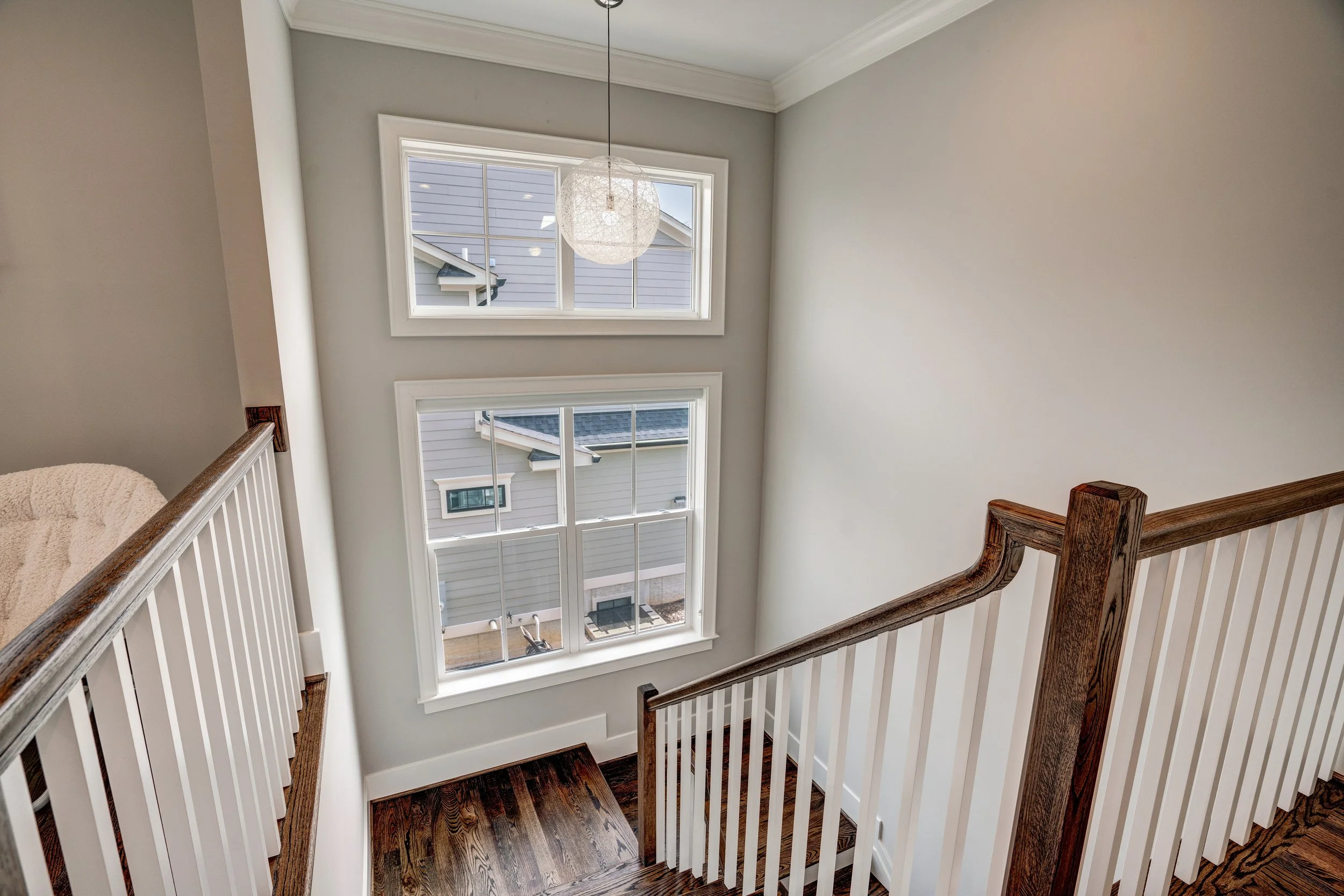 A view of a staircase landing with wooden handrails and white balusters, overlooking large windows showing neighboring houses with gray siding and black roofs, and a decorative light fixture hanging from the ceiling.