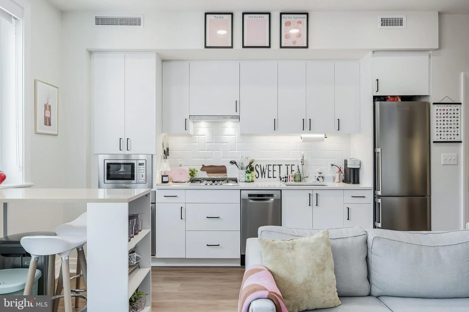 Modern white kitchen with stainless steel appliances, open shelving, decorative artwork, and a cozy living area in the foreground.