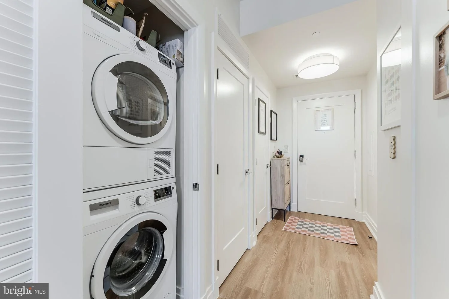 Laundry area with stacked washer and dryer, white walls, wooden flooring, small dresser, and front door with a pink checkered rug.