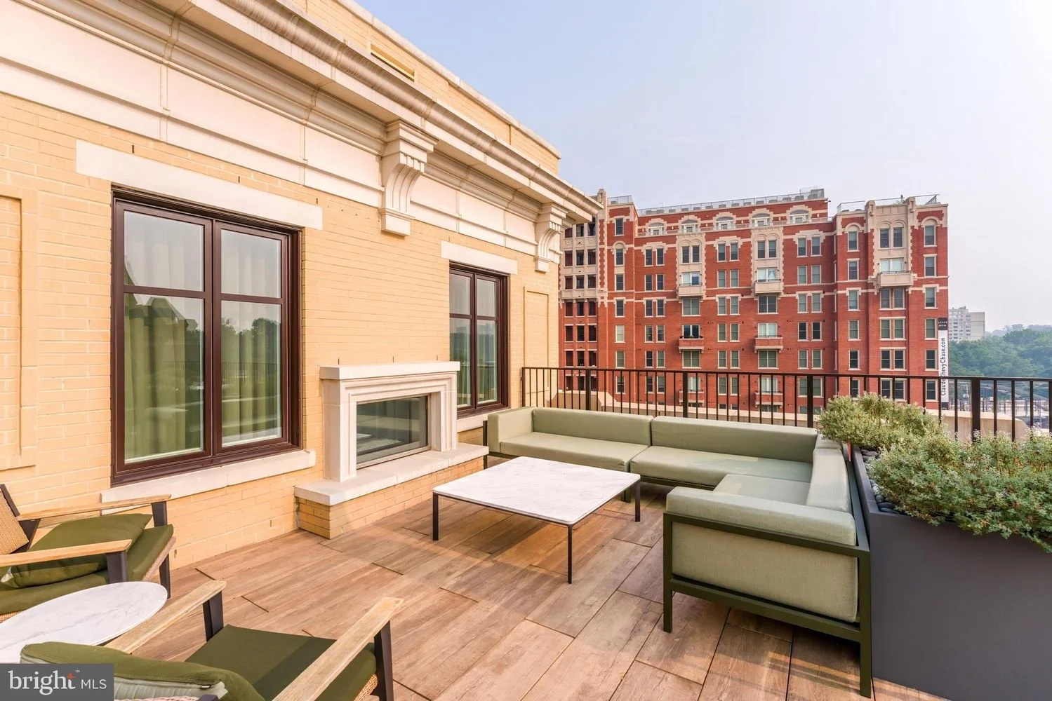 City balcony with green sectional sofa, matching chairs, a square marble-top coffee table, potted plants, and a view of a red brick building and blue sky.